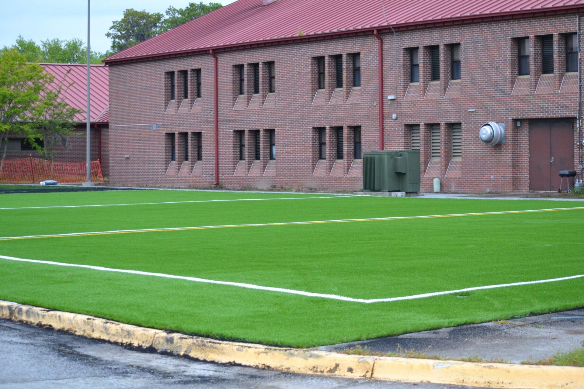 An artificial green turf field with white markings next to a red-brick building with metal barred windows and a red-tiled roof on an overcast day.