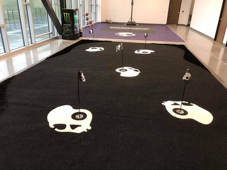 This image shows an indoor miniature golf course with carpet flooring featuring holes marked by flagsticks and skull-and-crossbones designs, and a vending machine on the left.