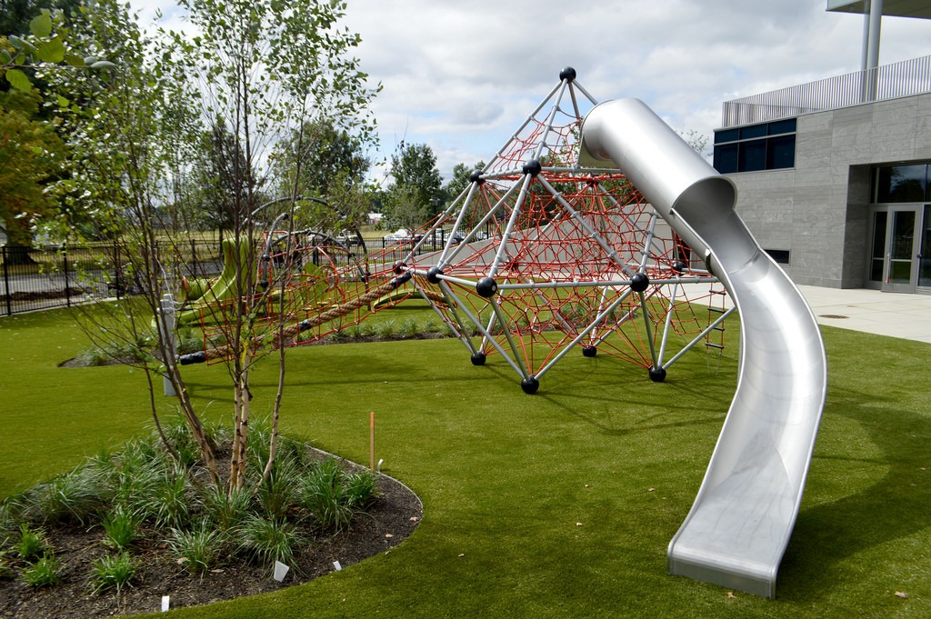 Modern playground with a geometric climbing structure and a tall, curved metal slide on artificial grass, surrounded by young trees and a building.