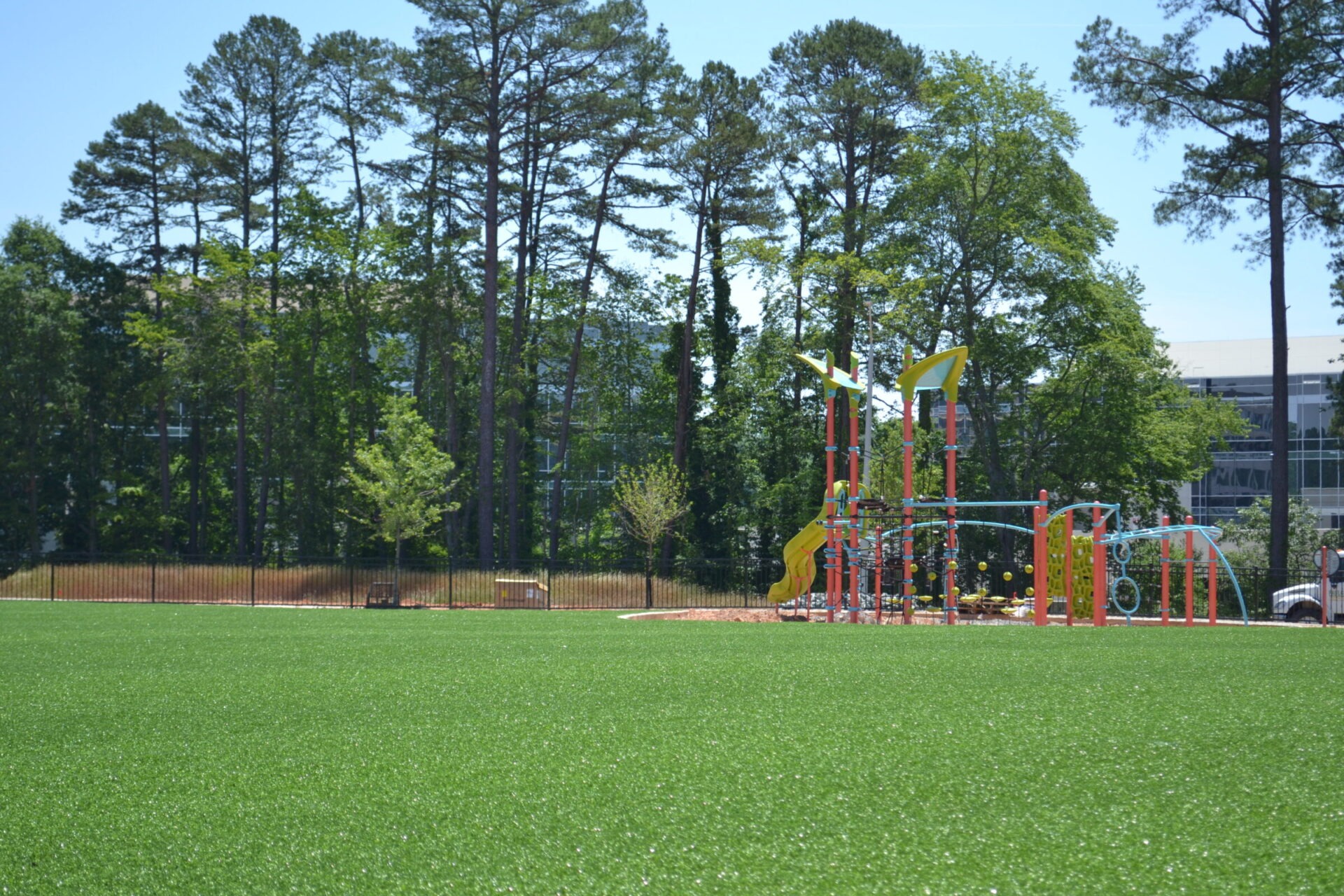 A playground with colorful equipment and slides set against a backdrop of tall trees and a clear blue sky, with an artificial turf foreground.
