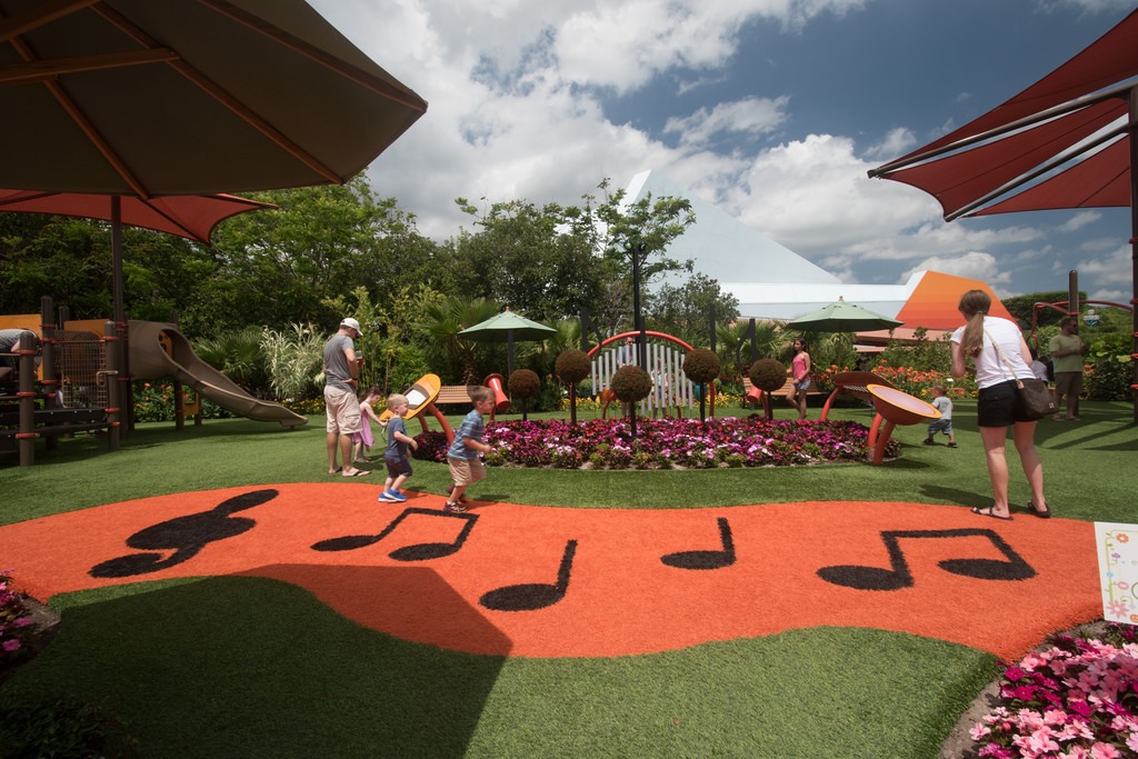 A vibrant outdoor playground with people, musical notes on the ground, slides, flowers, and shade umbrellas under a partly cloudy sky.