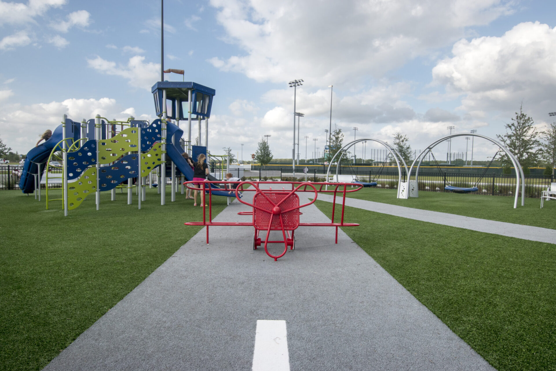 An outdoor playground featuring a red table, climbing structures, artificial turf, and people engaging in various activities under a partly cloudy sky.