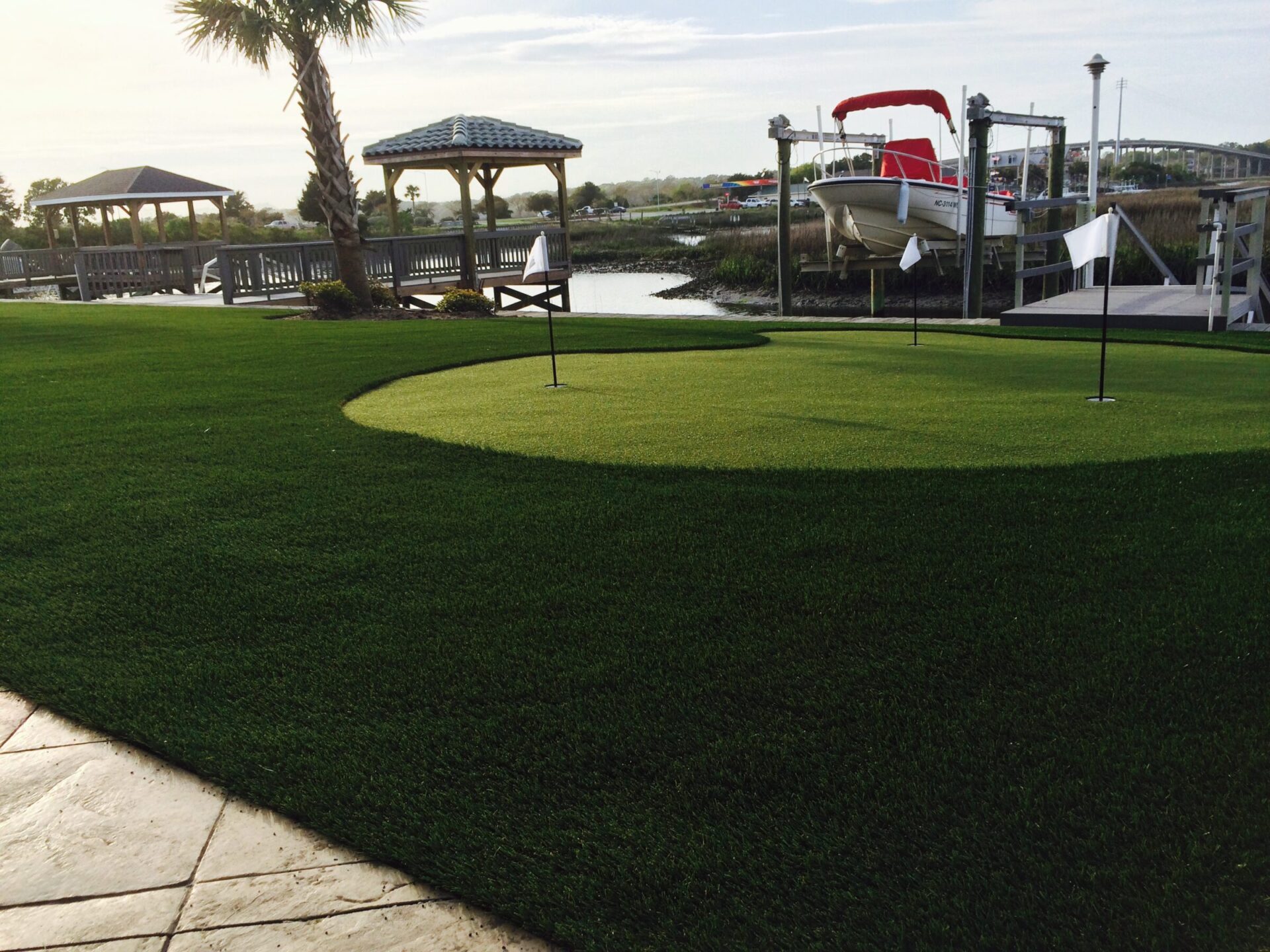 A manicured green lawn with putting flags beside a tranquil waterfront setting, featuring a boat on a lift and a gazebo under clear skies.