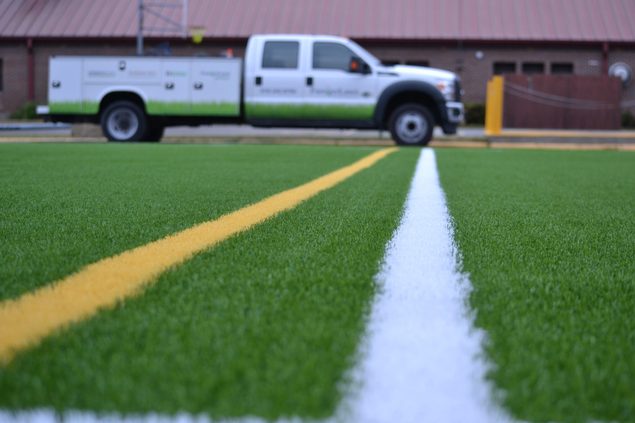 This image shows a close-up of white and yellow lines on lush green artificial turf, with a utility vehicle blurred in the background.