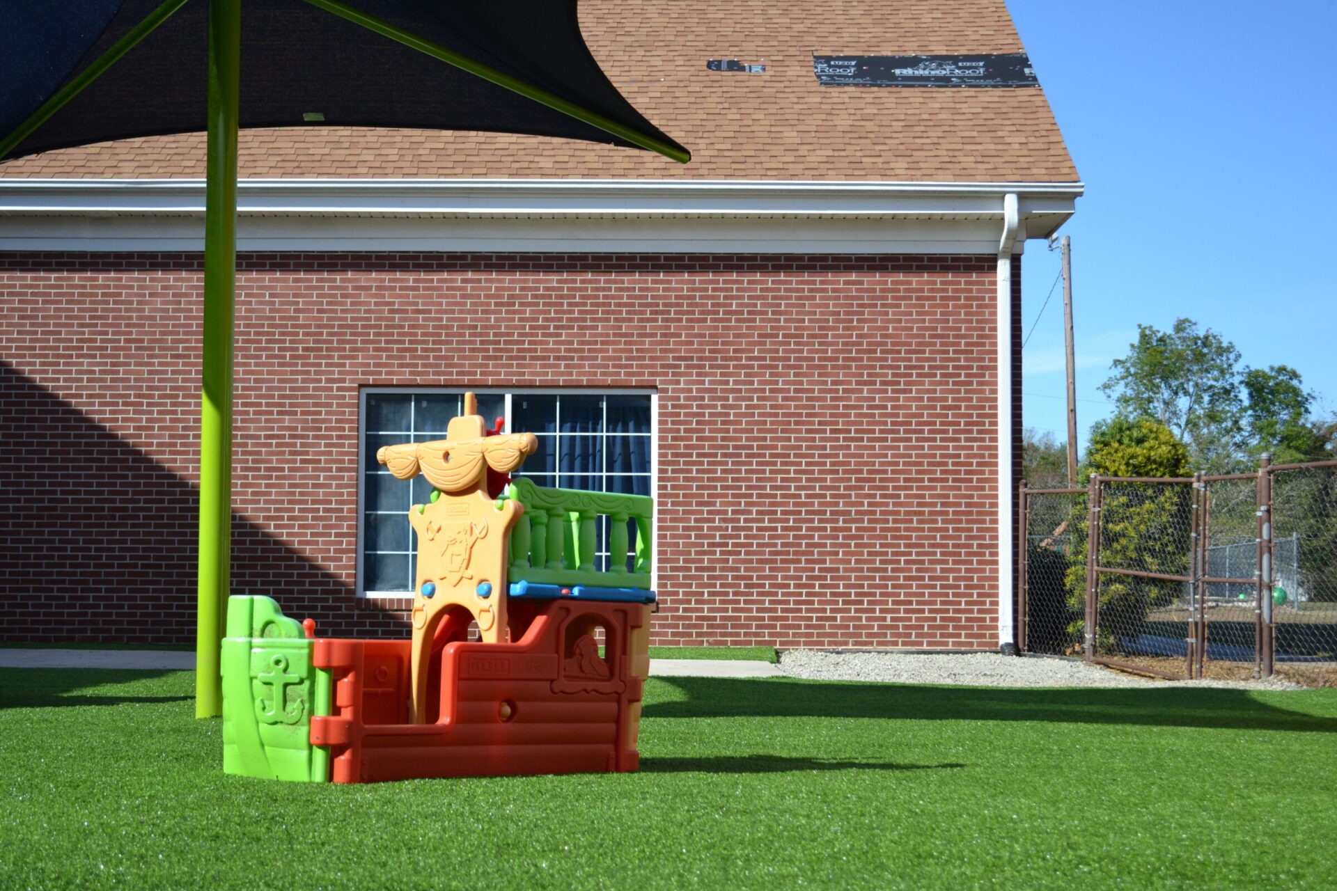 A colorful children's playground set stands on artificial turf, with a shaded canopy and a brick building in the background on a sunny day.
