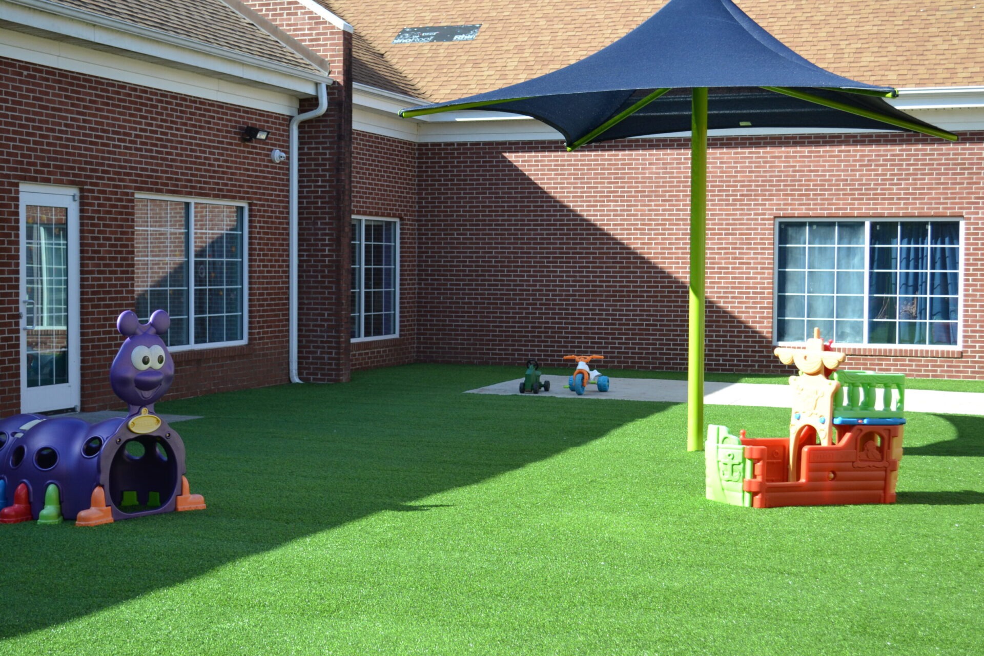 A playground with artificial grass features colorful play structures, including a purple caterpillar and a sunshade, against a brick building backdrop. No people visible.