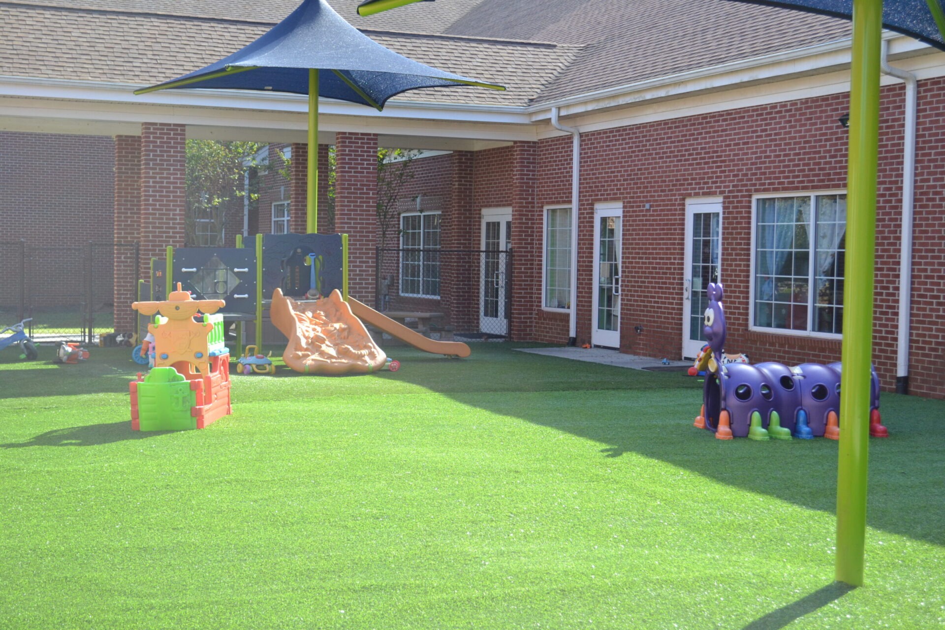 This is a brightly lit playground with colorful equipment, artificial grass, and a red brick building in the background, underneath a clear blue sky.