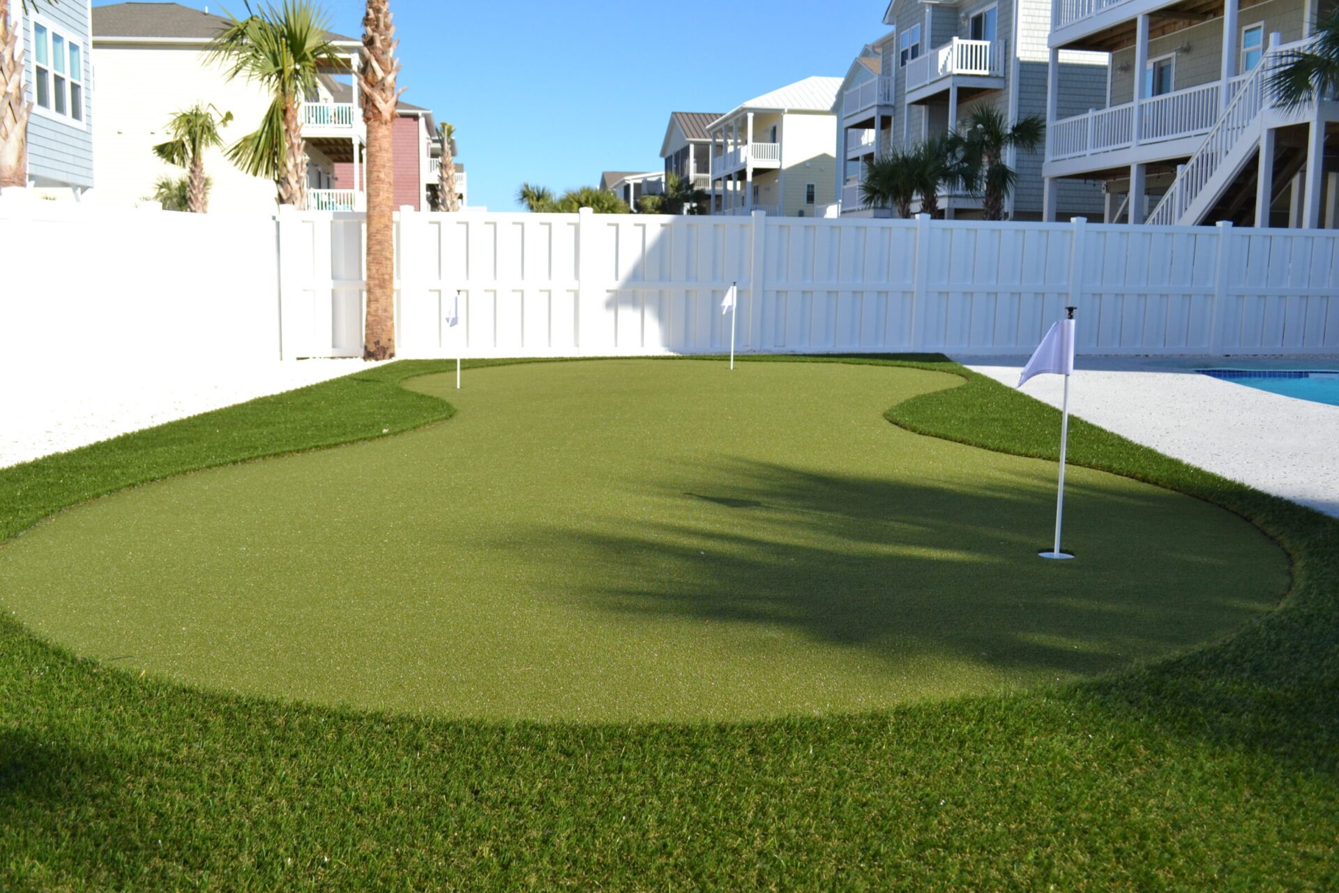 This is a backyard with a synthetic putting green next to a swimming pool, surrounded by a white fence and some beach houses with palm trees.