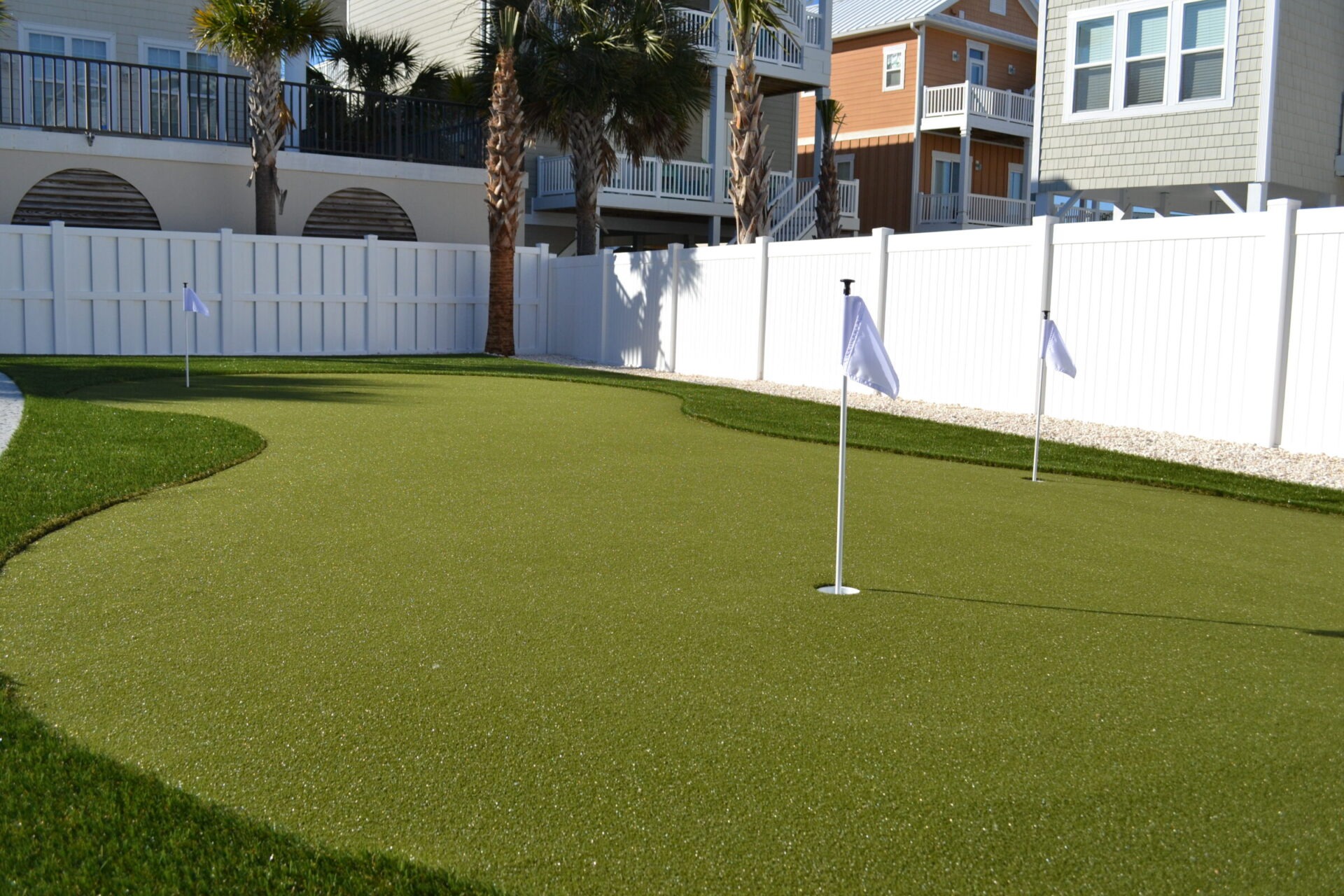 A backyard putting green with two holes and flags, surrounded by a white fence, palm trees, and residential buildings under a clear sky.