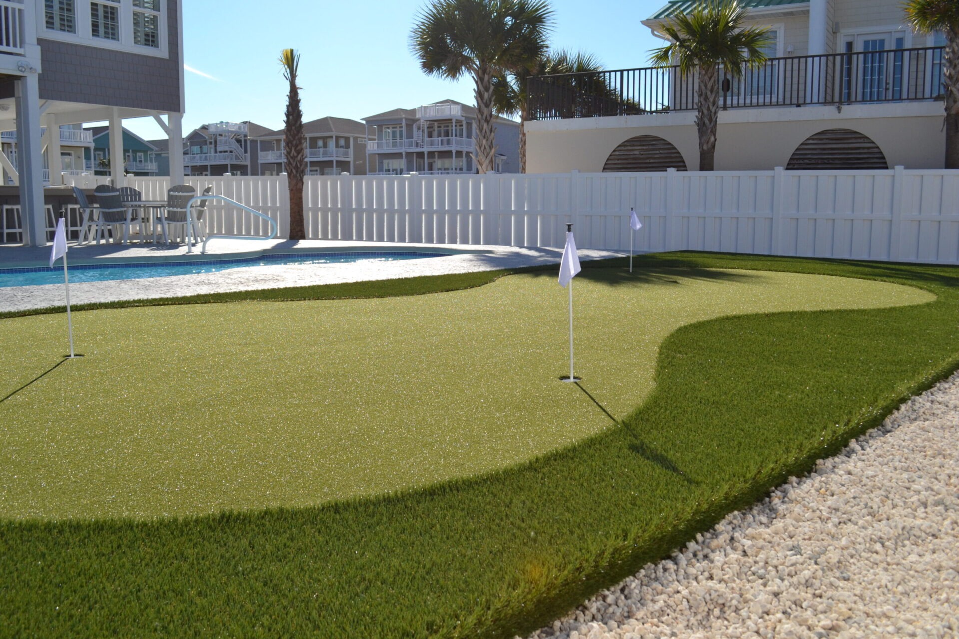 This image shows an artificial turf putting green with two golf holes and flags, near a pool and white-fenced residential buildings on a sunny day.
