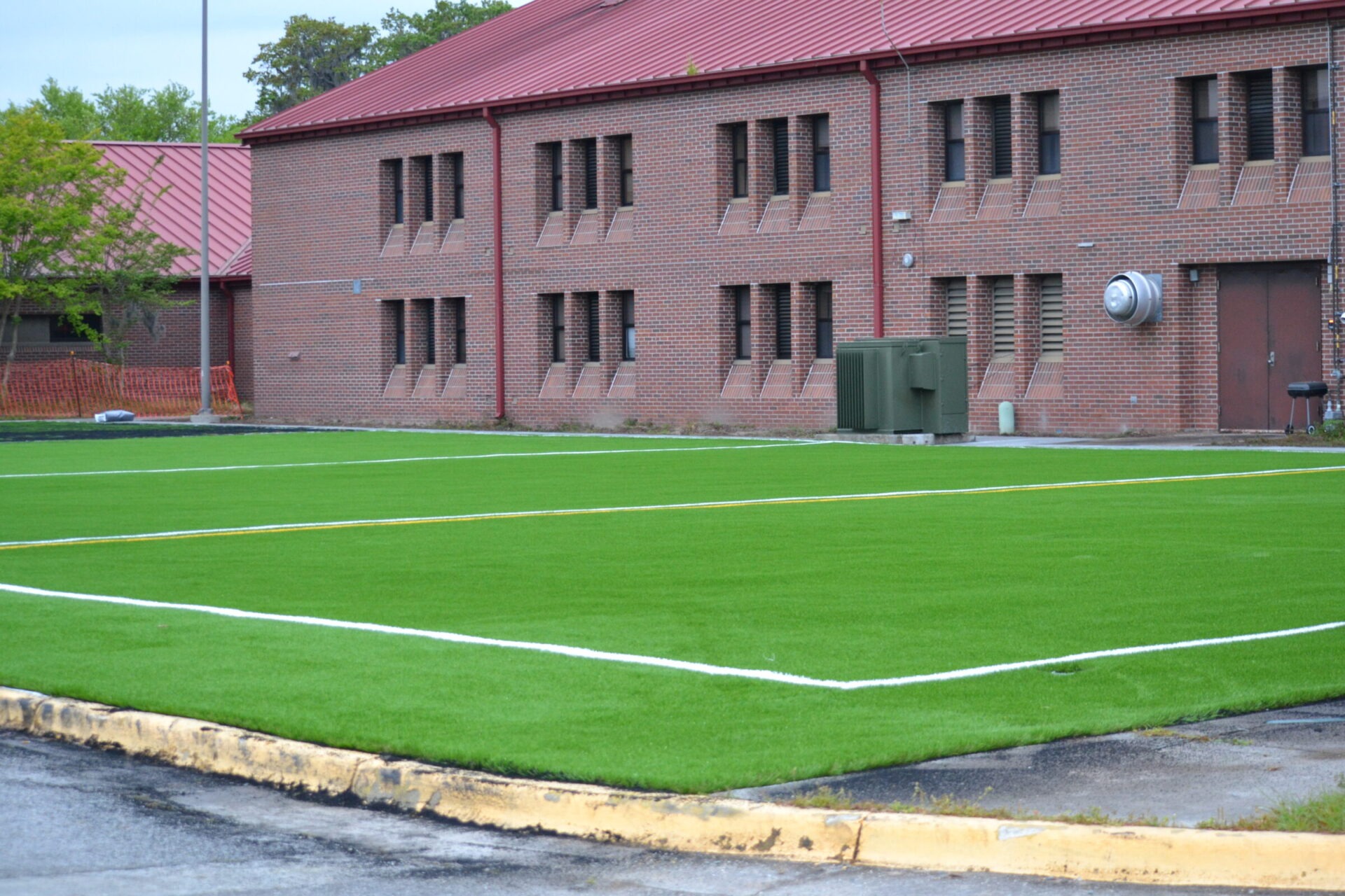 A newly installed artificial turf field is adjacent to a brick building with multiple windows and some utility equipment outside. The weather appears overcast.