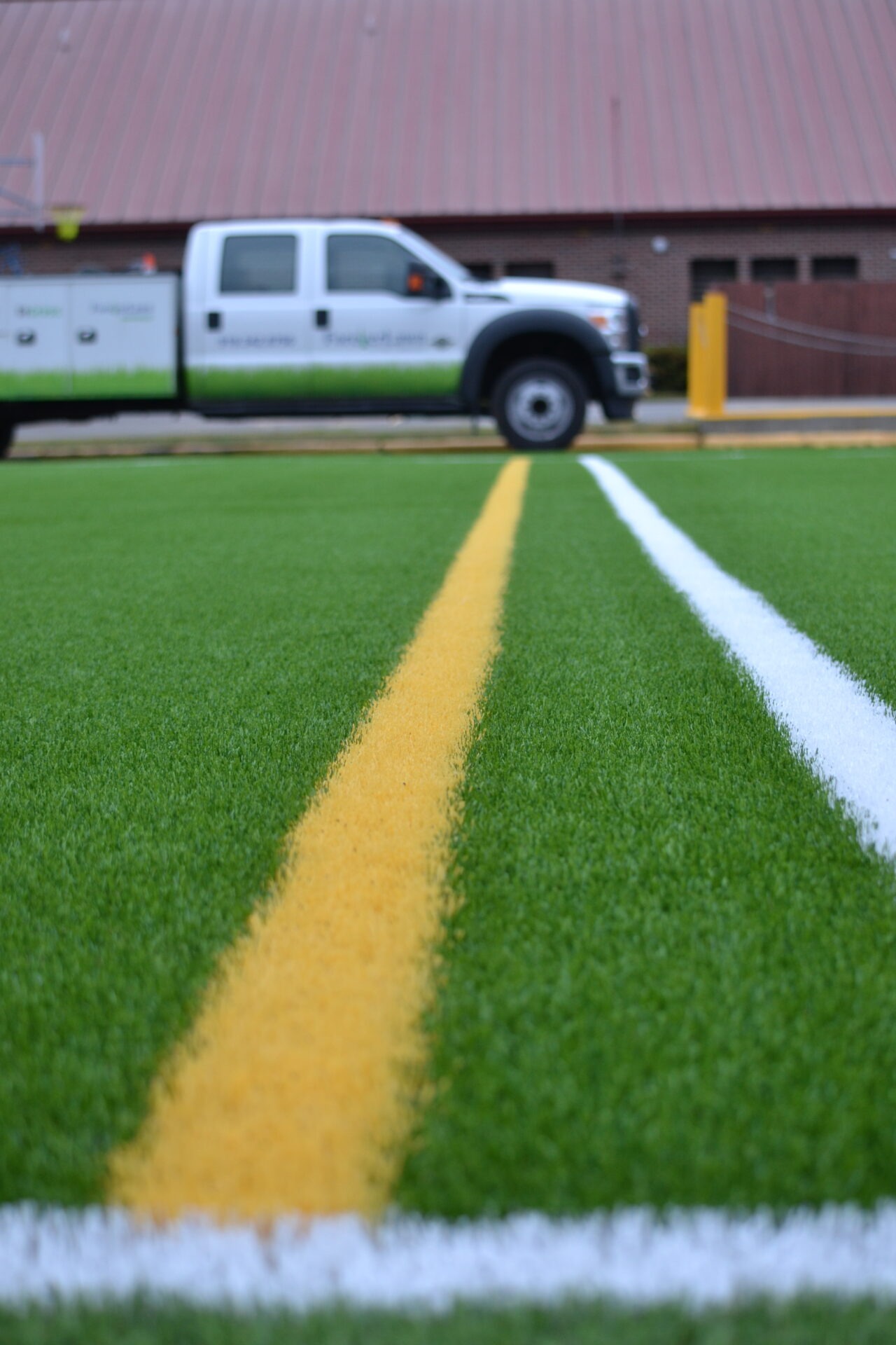 The image features a close-up view of artificial green turf with yellow and white boundary lines. In the blurred background, there's a parked vehicle.