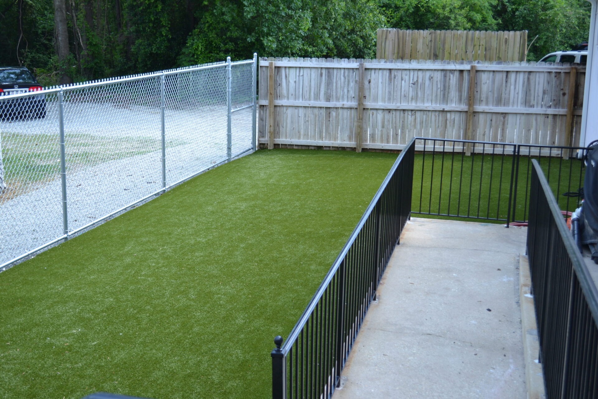 This image shows a fenced backyard with artificial green grass, a concrete path, a chain-link fence, and a wooden privacy fence.