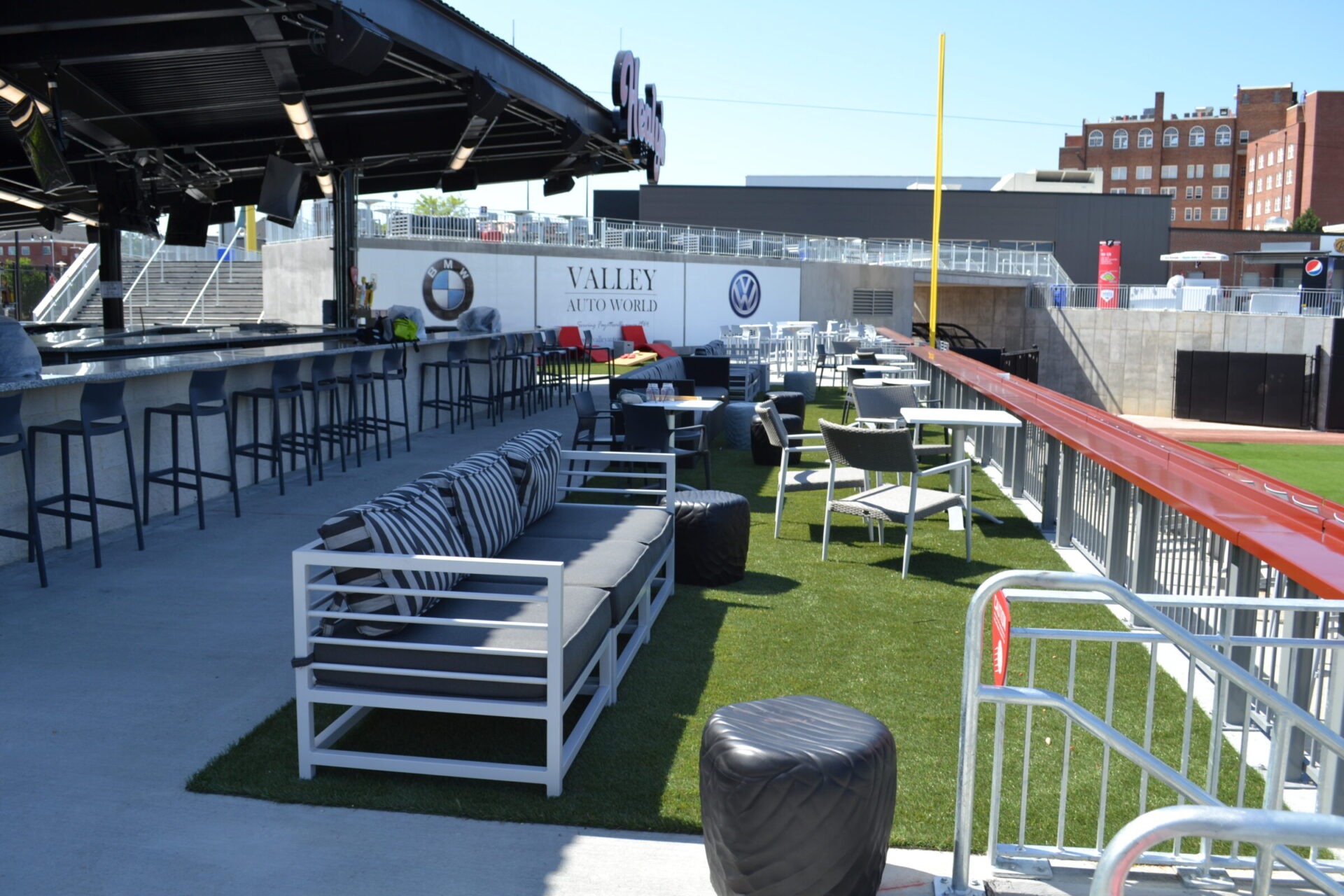 This image shows an outdoor lounge area with modern furniture, artificial grass, a bar, and a clear view of a sports field's foul pole.