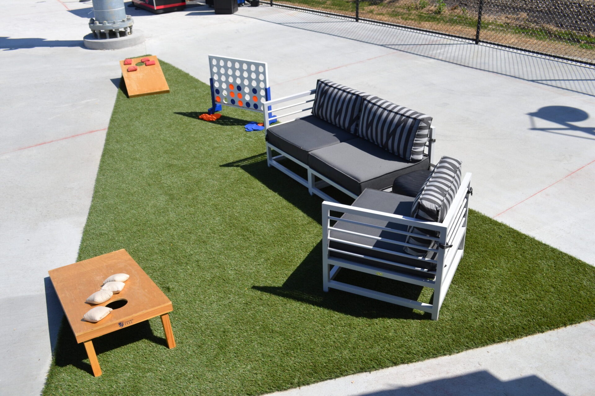An outdoor recreational area with artificial turf, featuring games like cornhole and Connect Four, a lounge set with striped pillows, and a small wooden table.