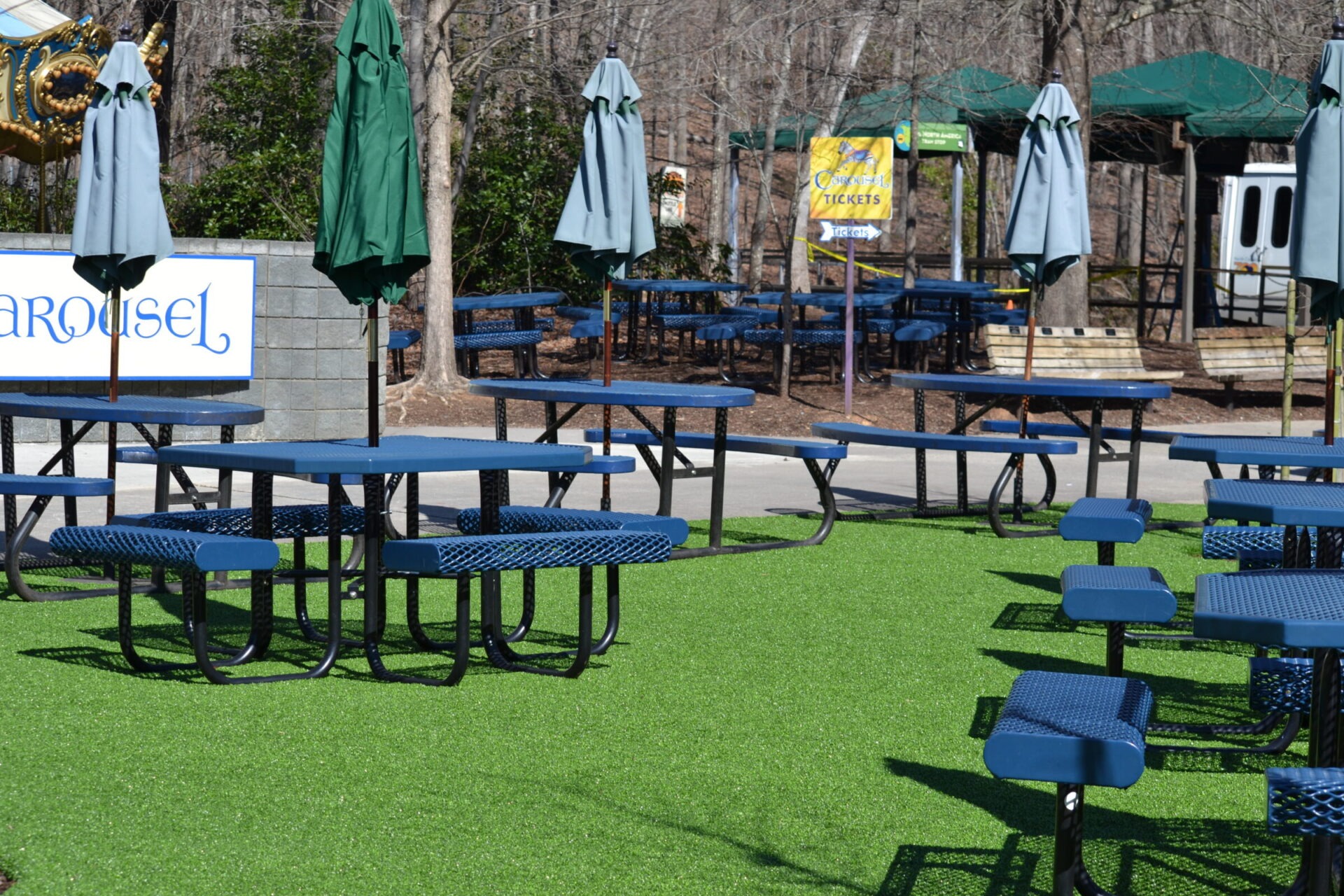 The image shows an outdoor seating area with blue picnic tables, closed green umbrellas, artificial grass, and signage for a carousel and tickets.