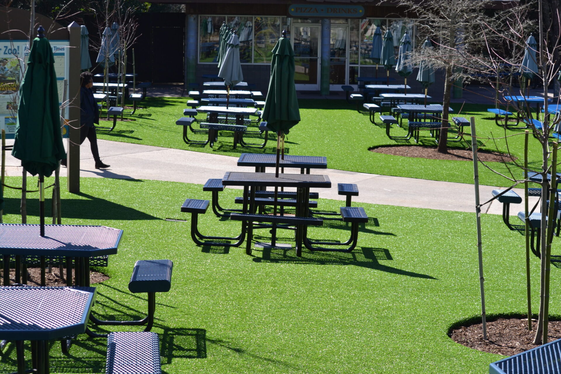 Outdoor seating area with green artificial turf, multiple picnic tables, closed umbrellas, and a person walking by under a clear, sunny sky.