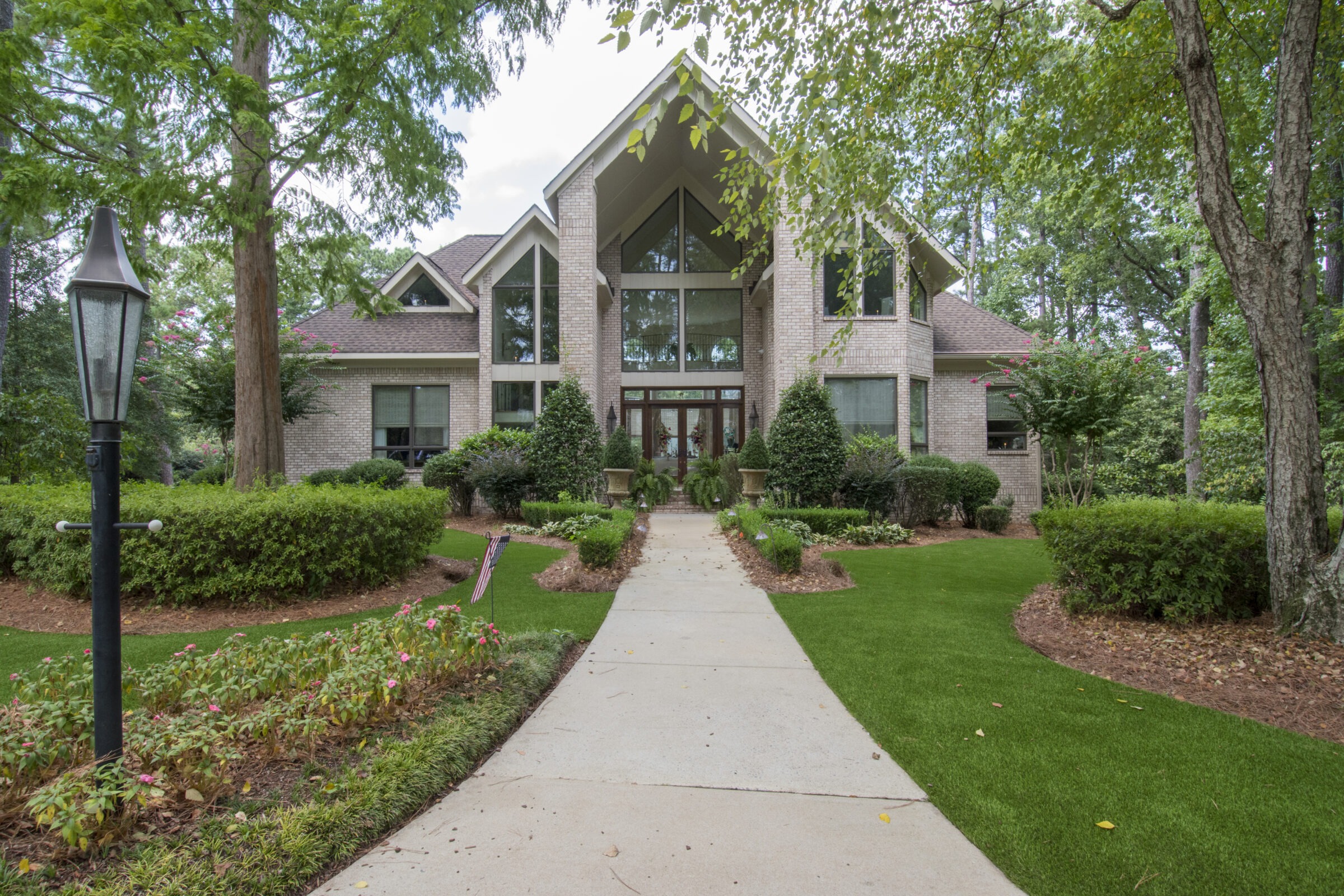 A large brick house with prominent gables and large windows, landscaped garden, concrete pathway, and an outdoor lamp post in the foreground.