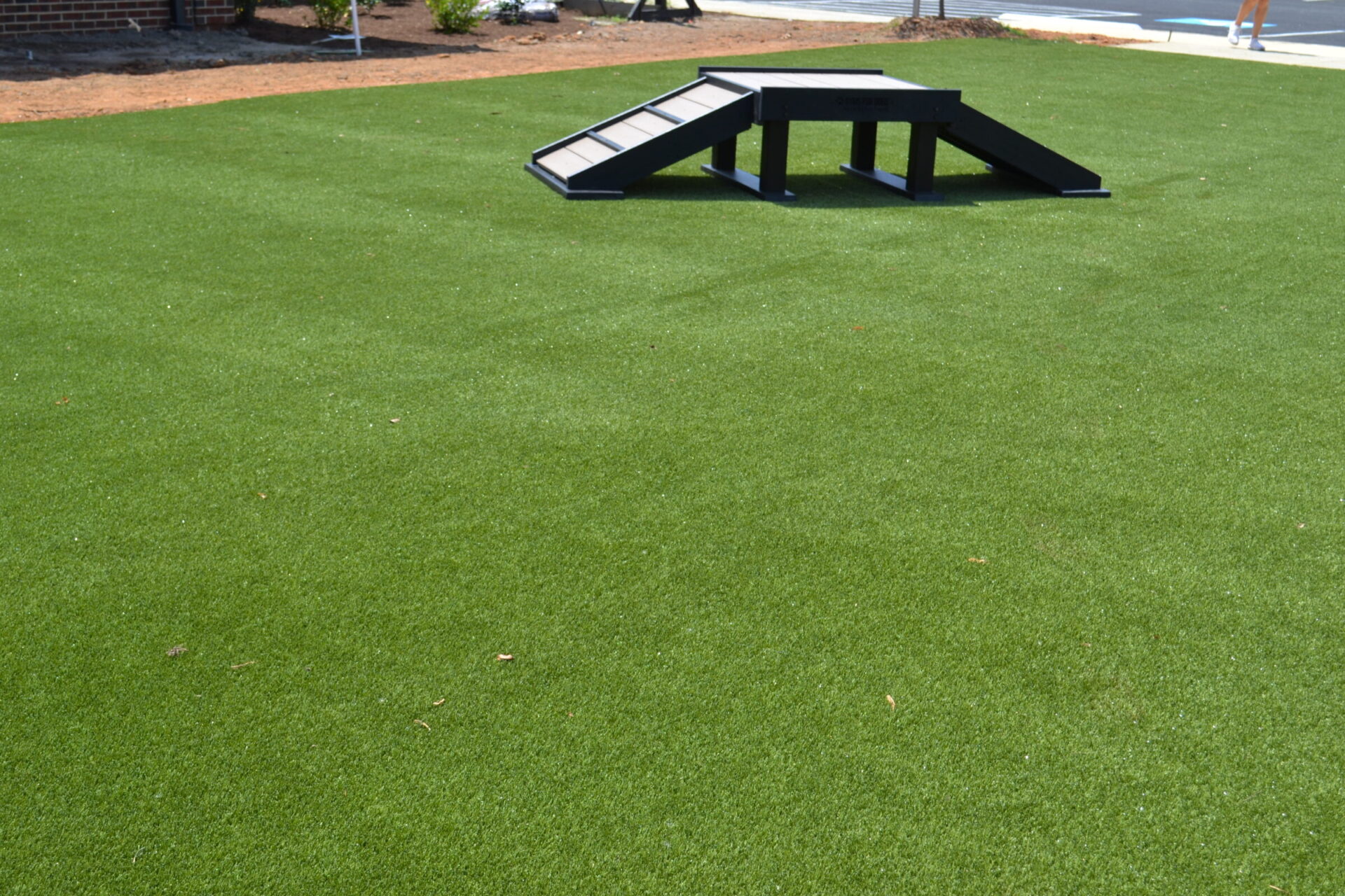 A black dog agility ramp sits on artificial green grass with a clear sky above and a person visible in the background on the right.