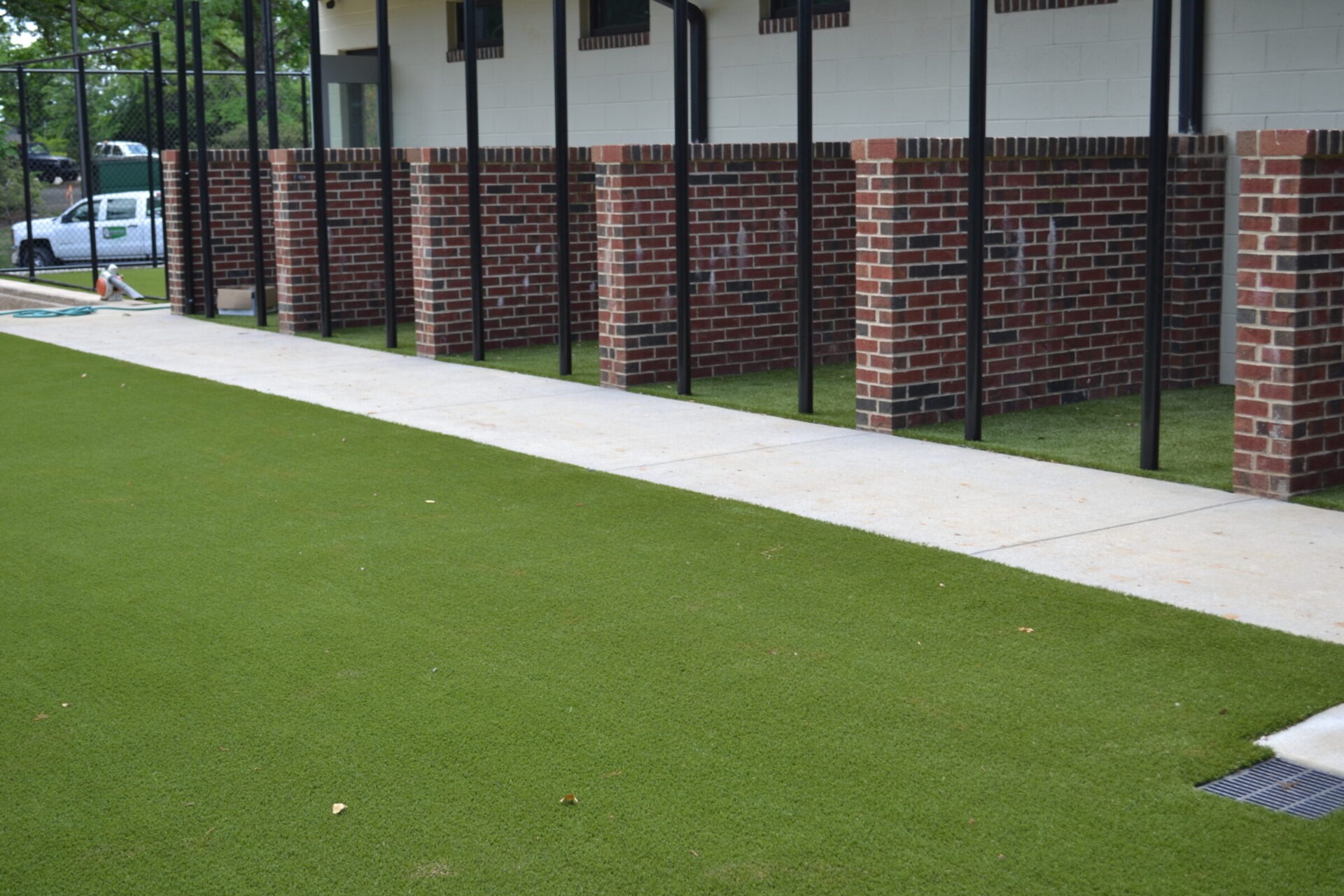 The image shows a well-maintained area with artificial green turf bordering a concrete walkway beside a brick structure with black metal fencing.