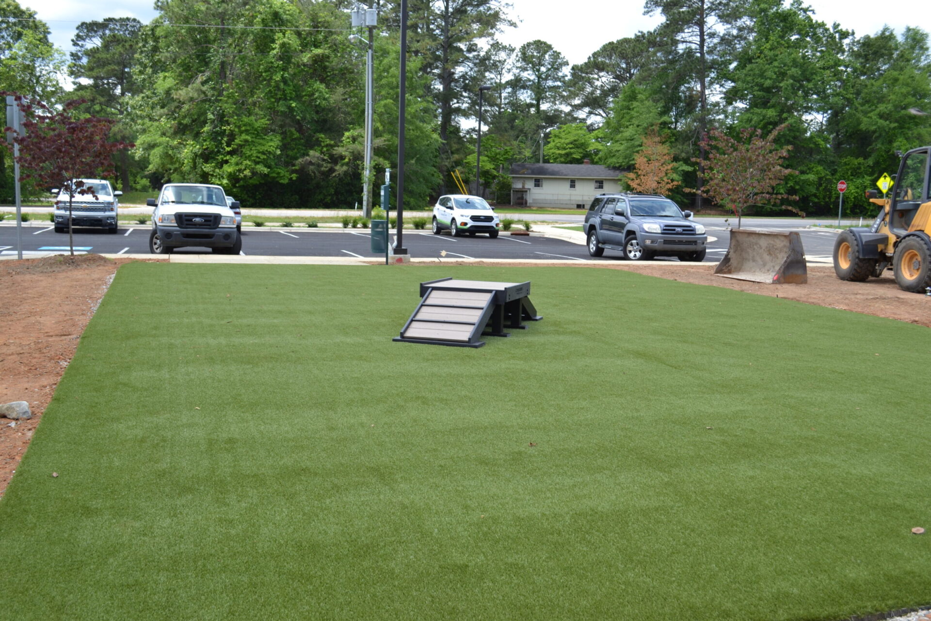 This image shows an artificial grass area with a black agility ramp for dogs, adjacent to a parking lot with vehicles and a backhoe.