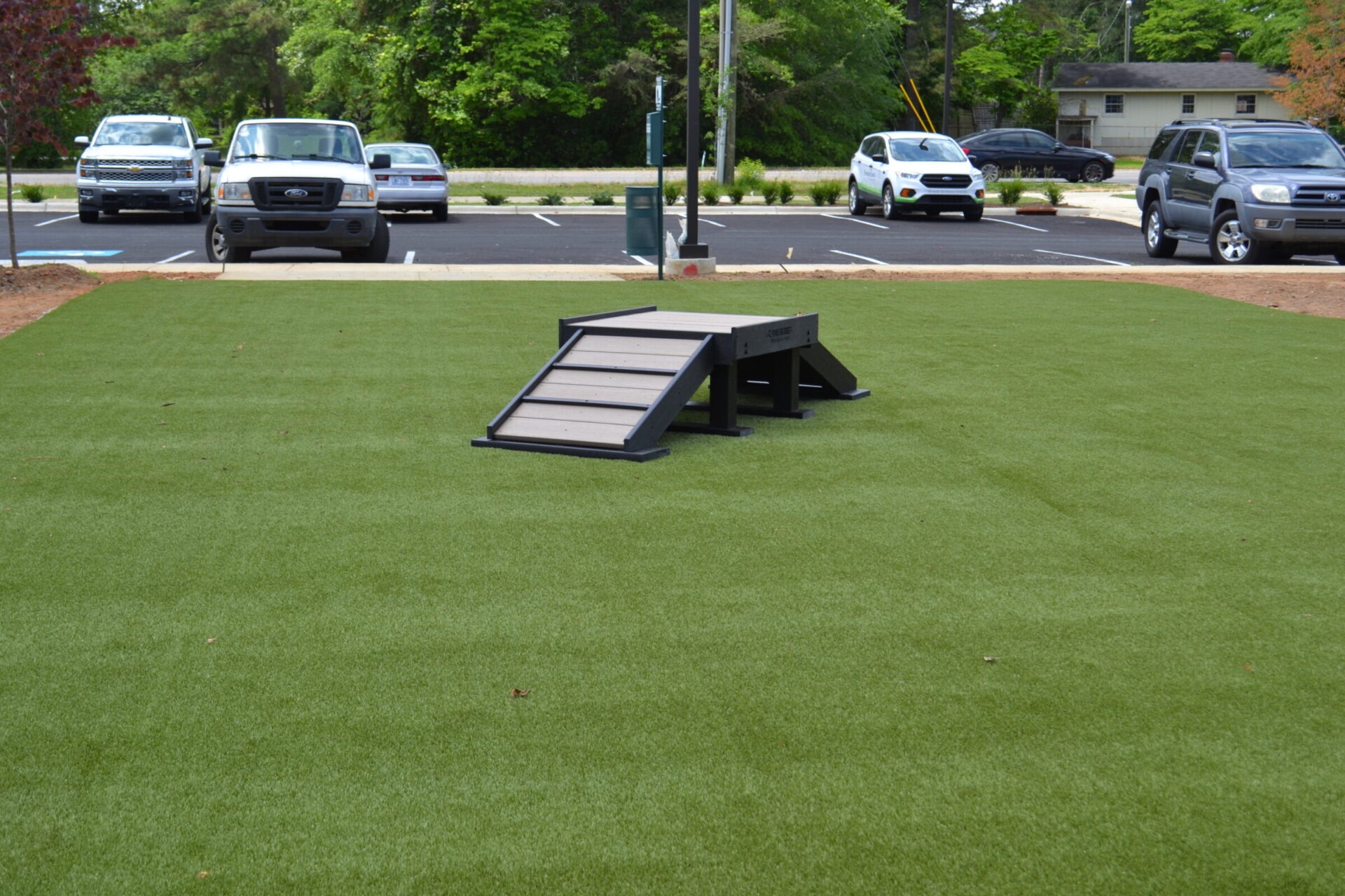 An agility ramp for dogs is on artificial grass with parked vehicles in the background and trees and a road further behind.