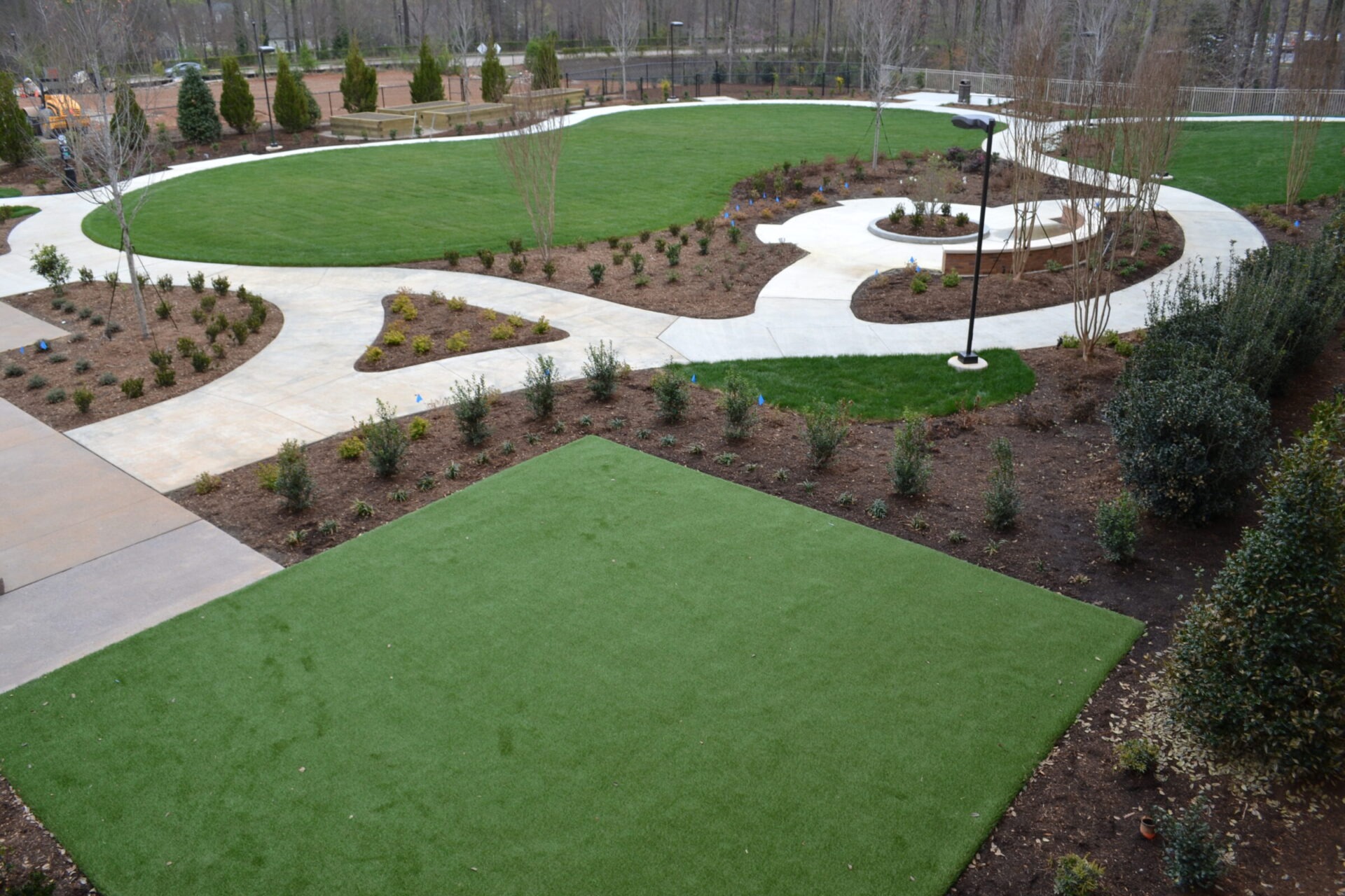 Newly landscaped park with curving pathways, green lawns, young trees, and mulched garden beds under a cloudy sky. A streetlamp stands beside the path.
