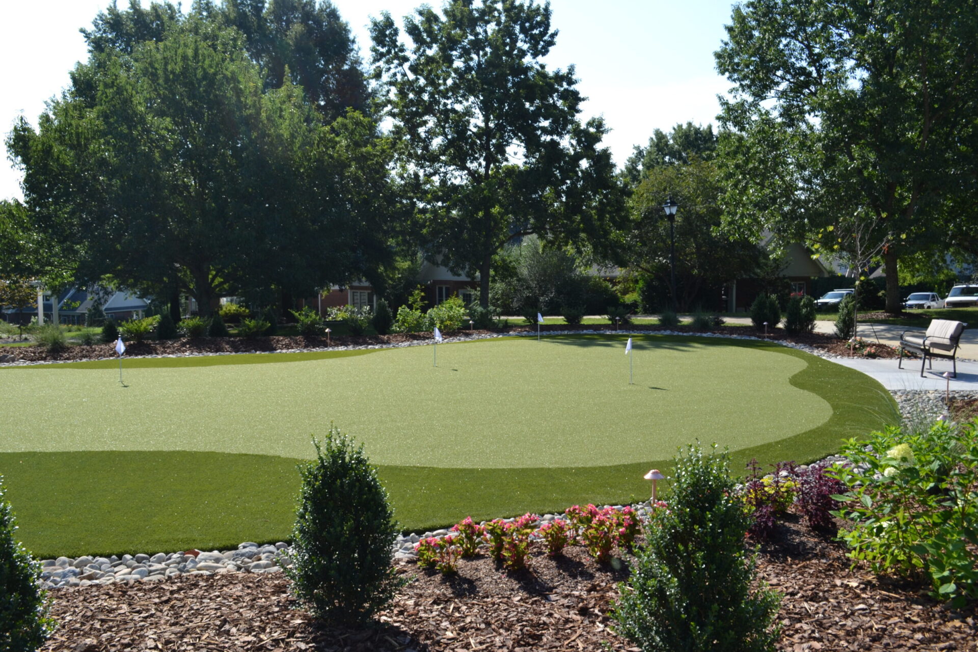 A well-manicured artificial putting green is surrounded by flowering plants, rocks, and trees in a sunny suburban garden setting with benches nearby.