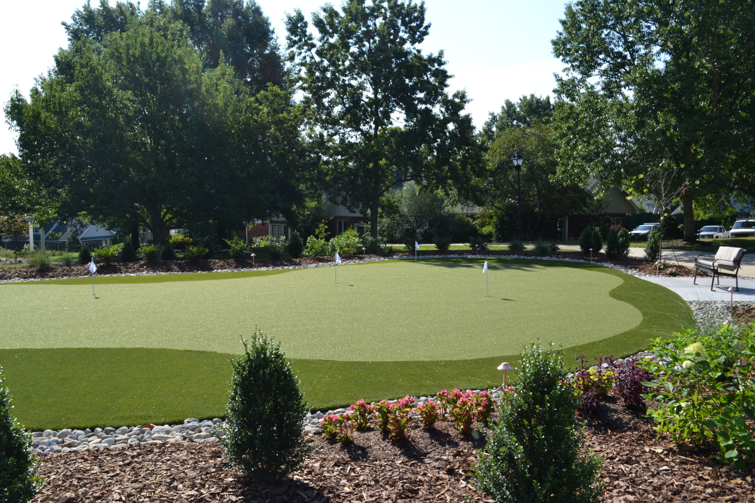 A well-manicured artificial putting green is surrounded by flowering plants, rocks, and trees in a sunny suburban garden setting with benches nearby.