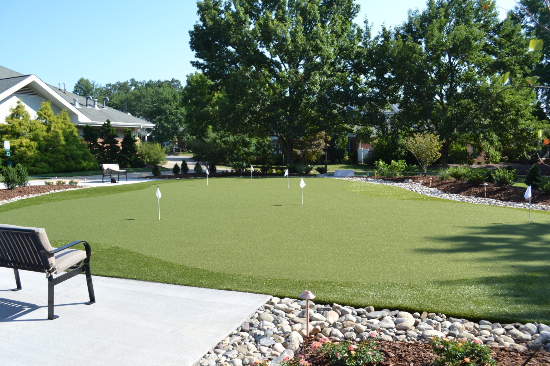 A well-maintained synthetic putting green with several holes and flags is surrounded by shrubs, trees, a bench, and a cobblestone border.