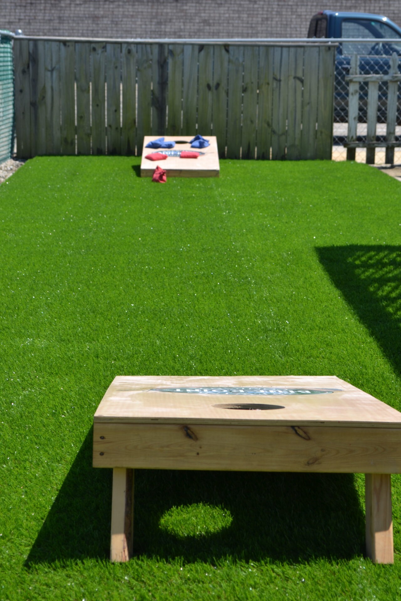 Two wooden cornhole game boards with red and blue bean bags are set up on artificial grass in a fenced backyard on a sunny day.