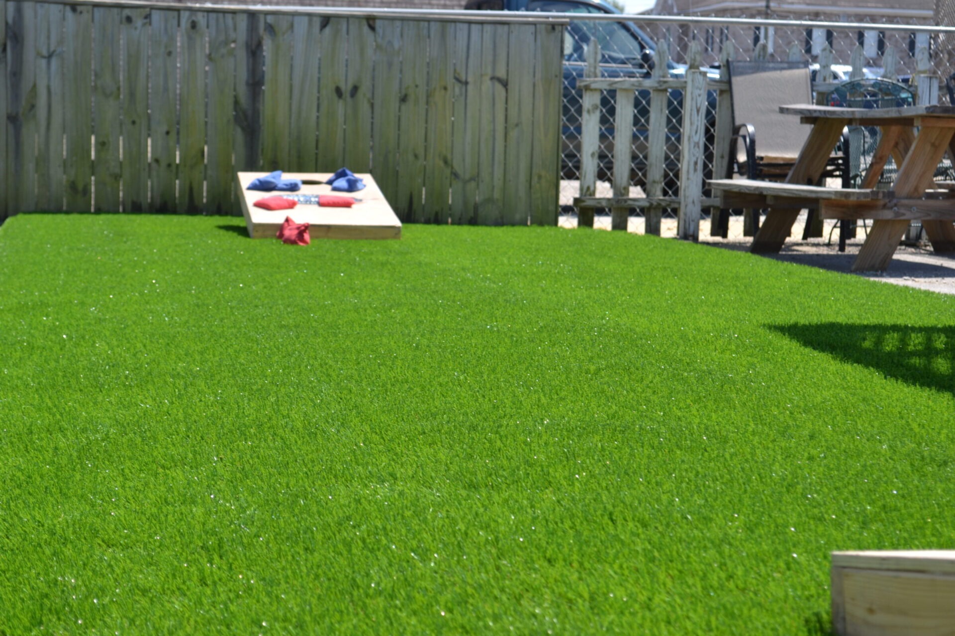 This image shows a vibrant green lawn with a cornhole game set up. Beanbags are on the boards and ground. A wooden picnic table is in the background.