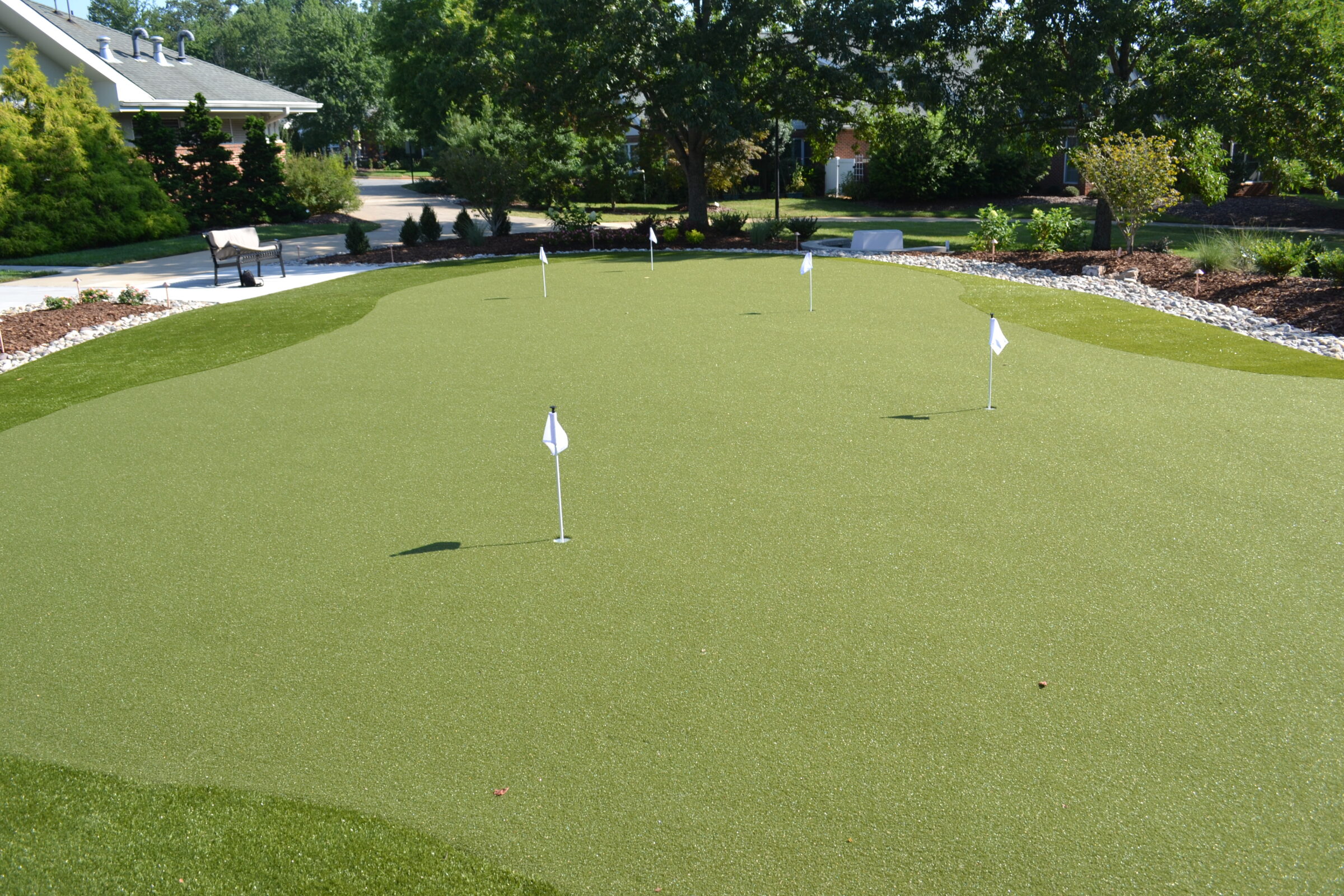The image depicts an artificial turf putting green with multiple golf holes and white flags, surrounded by landscaped garden and benches.