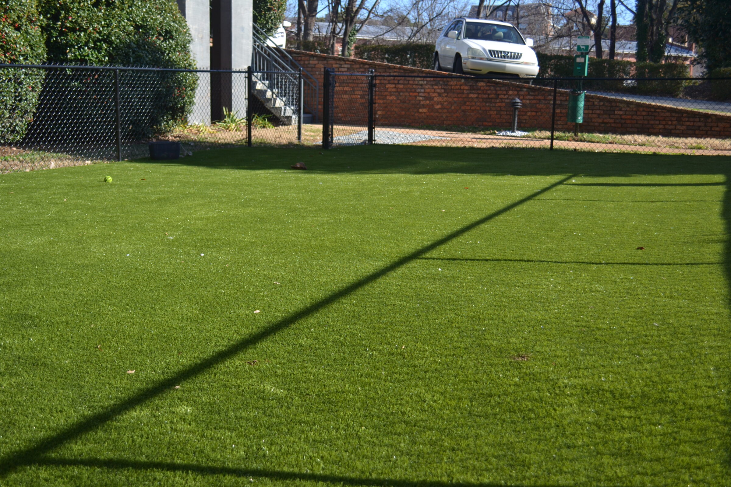 Lush green artificial turf with shadows cast by a fence, a lone tennis ball visible, adjacent to a brick wall with parked car above.