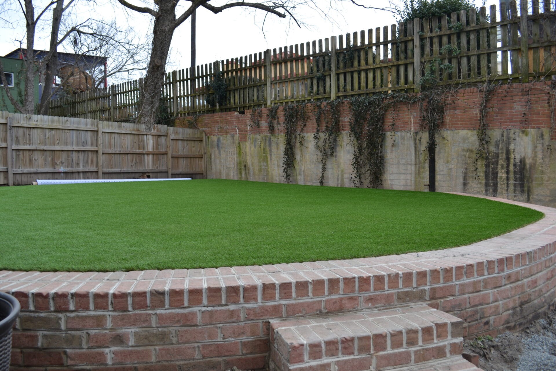 An outdoor space features a lush green lawn encircled by a curved brick retaining wall, with a wooden fence and vines visible in the background.