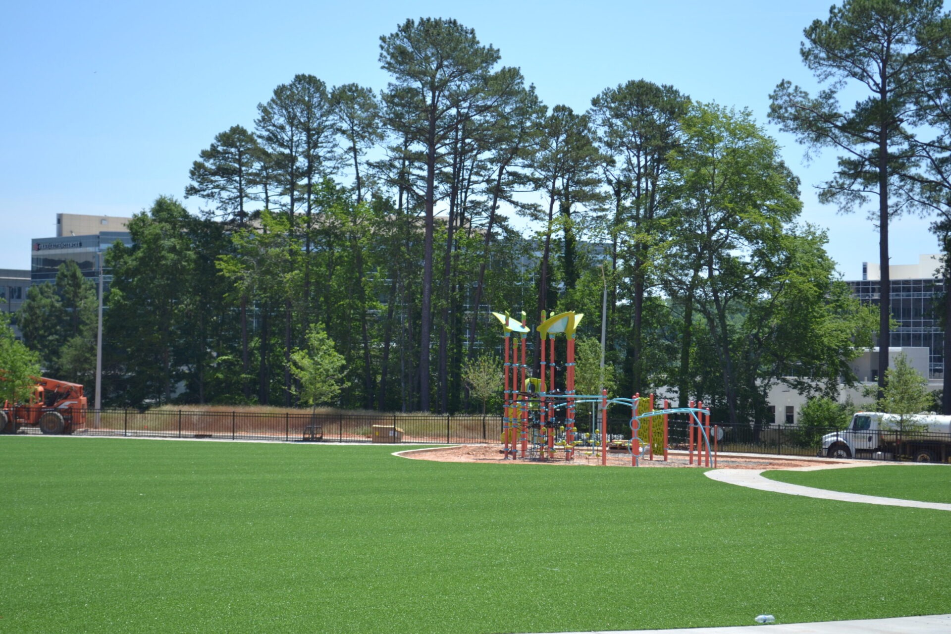 A vibrant playground with slides and climbing structures sits on artificial grass, surrounded by tall pine trees, under a clear blue sky.