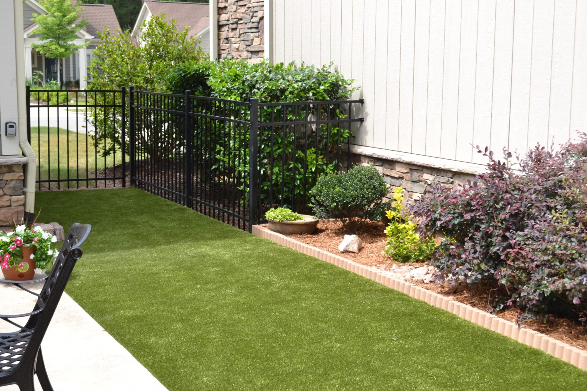 This image shows a well-manicured garden with artificial grass, a black iron fence, varied shrubs, and a stone and siding house facade.