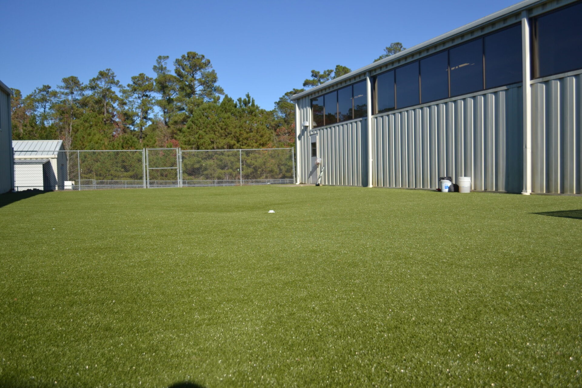 An expansive artificial turf field beside a metal-clad building with large windows, edged by a chain-link fence, under a clear blue sky.