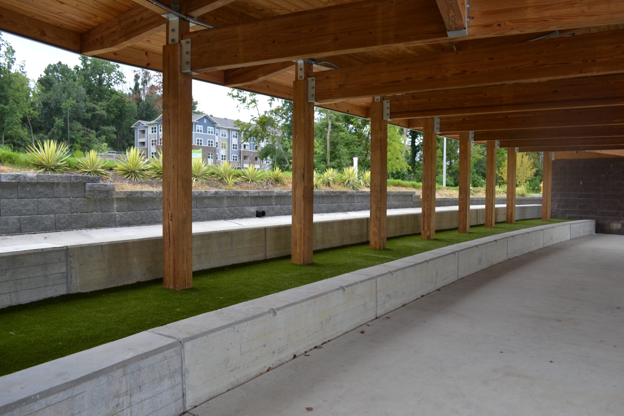 A covered outdoor area with wooden pillars supporting a roof, concrete seating, artificial grass, and landscaped beds with green plants and a multi-story building in the background.