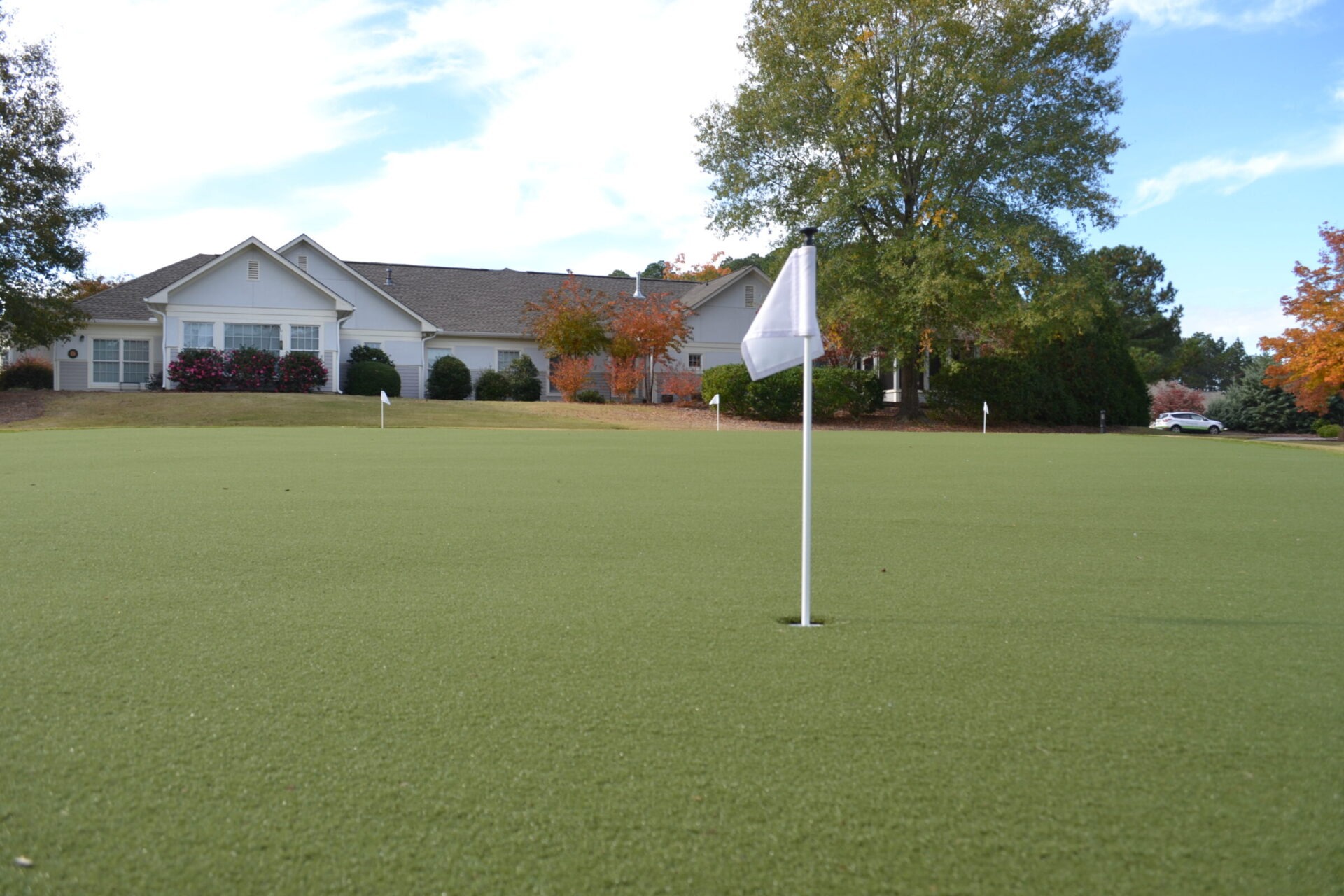 This is a photo of a golf green with a white flag in the hole, a background with a house, trees exhibiting autumn colors, and a car.