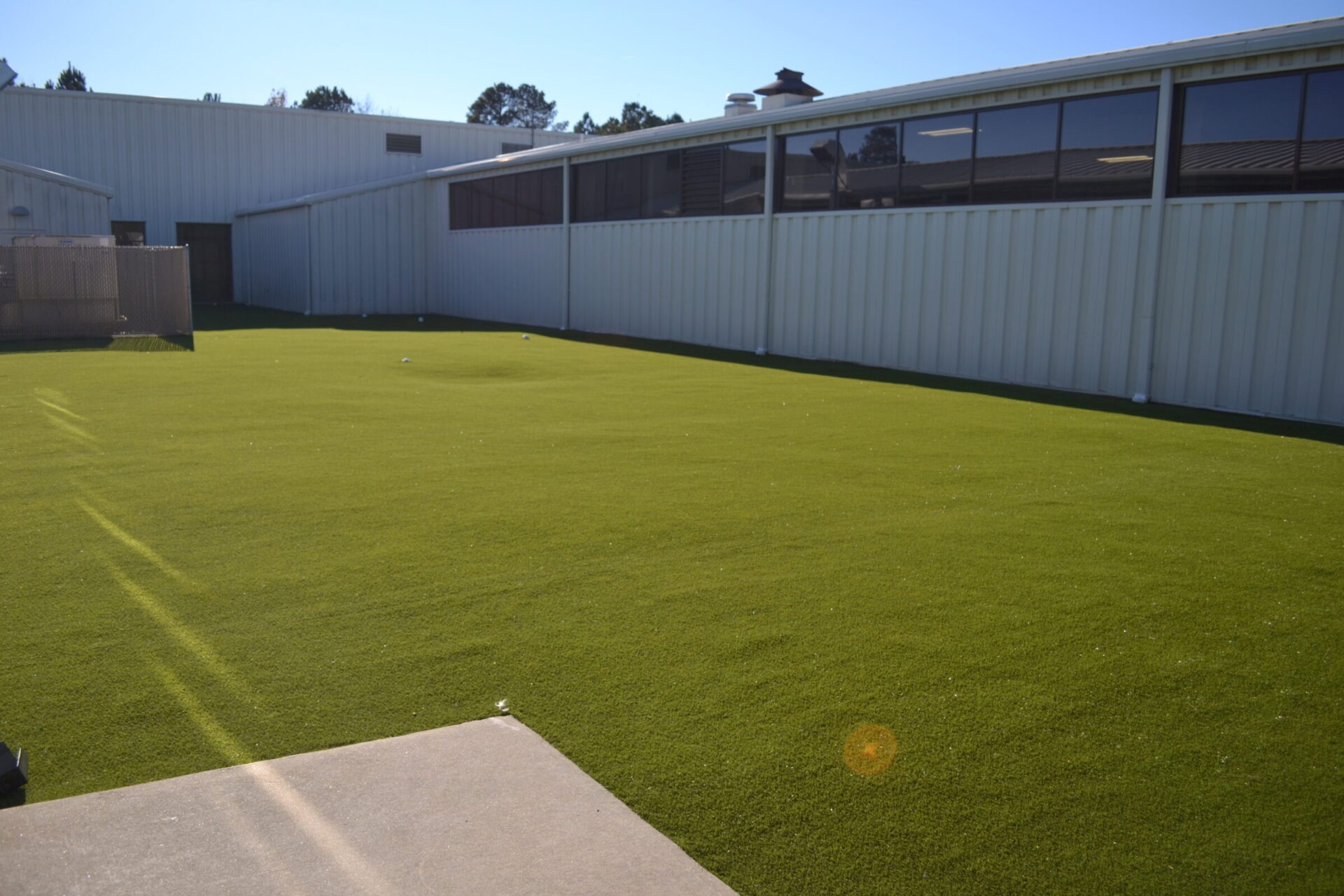 This image shows a large outdoor area covered with green artificial turf, bordered by a white industrial building with numerous windows under a clear sky.