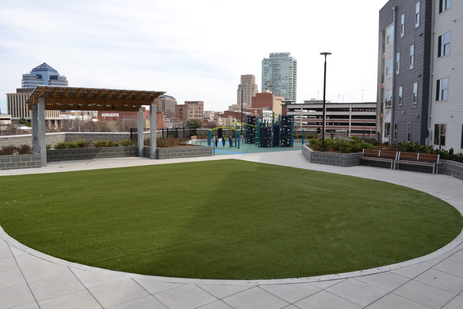 This image depicts a modern urban park with green artificial turf, a playground, seating areas, pergolas, and city buildings in the background.