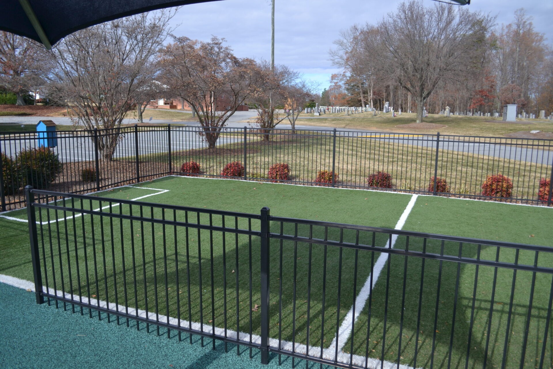 An outdoor view showing a fenced artificial turf area with lines resembling a mini sports field, deciduous trees, and a cemetery in the background.