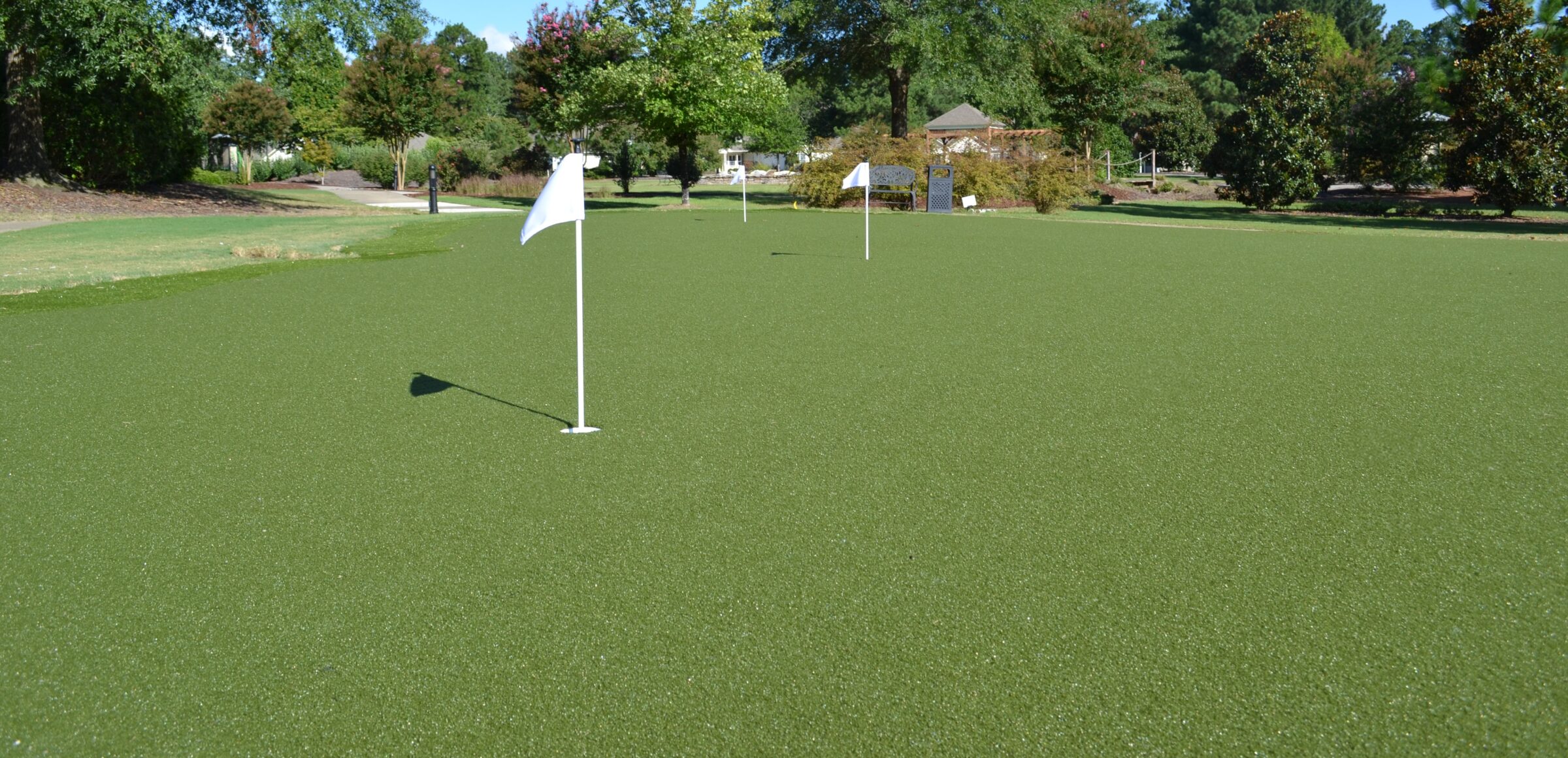 This is a wide-angle photo featuring a large, smooth artificial turf putting green with multiple holes marked by flags. Surrounding trees and clear skies are visible.