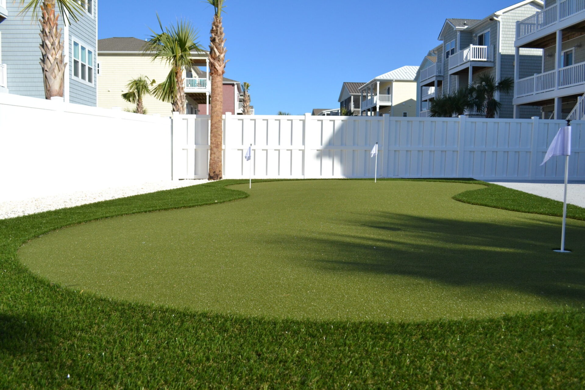 A small artificial putting green with holes and flags enclosed by a white fence, surrounded by houses and palm trees under a clear blue sky.