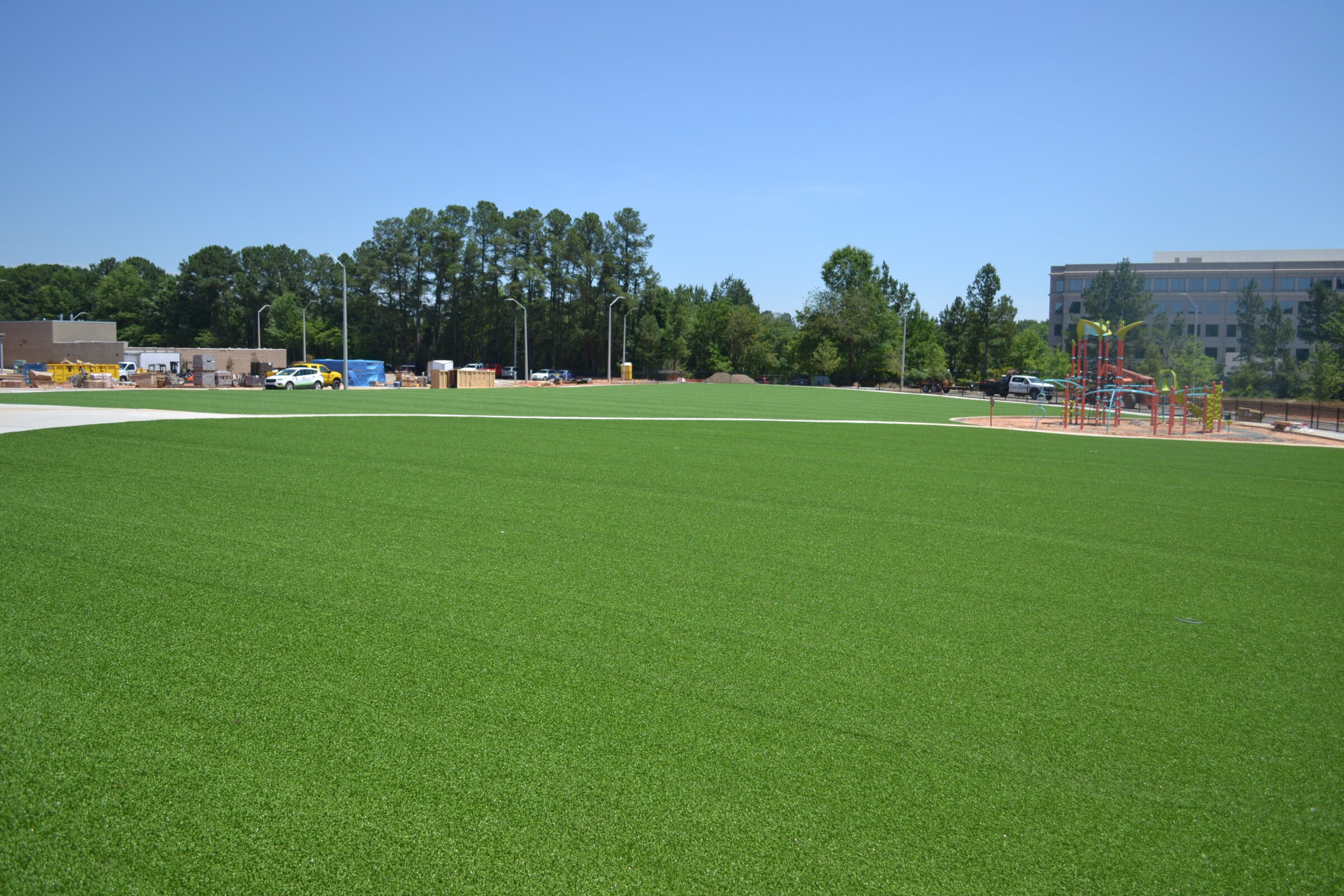A vibrant artificial turf field, clear skies, bordering woodland, playground equipment in the distance, with urban buildings and parked cars observable.