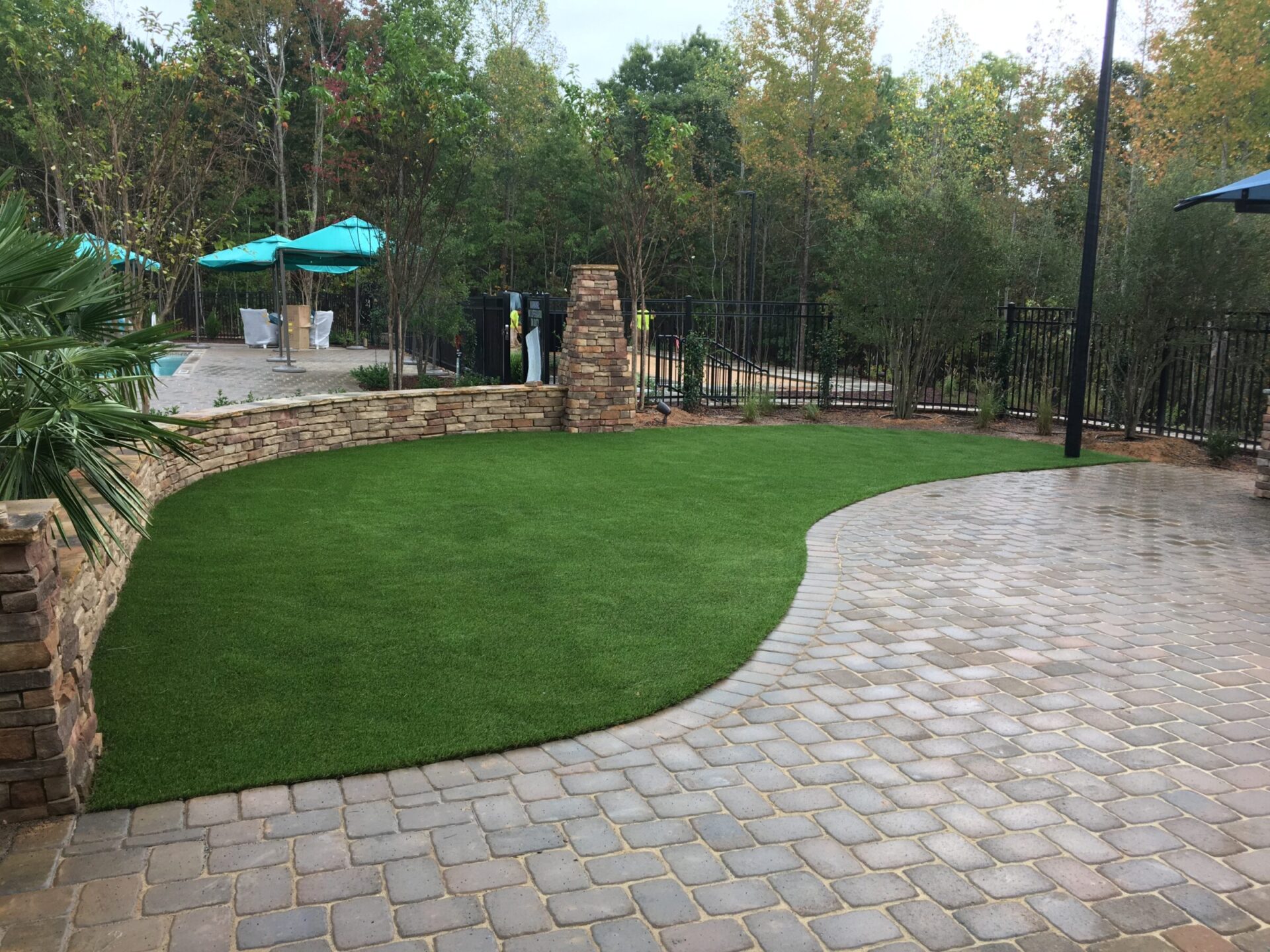 The image shows a landscaped area with artificial grass, stone paving, a curved retaining wall, outdoor umbrellas, trees, and a metal fence.
