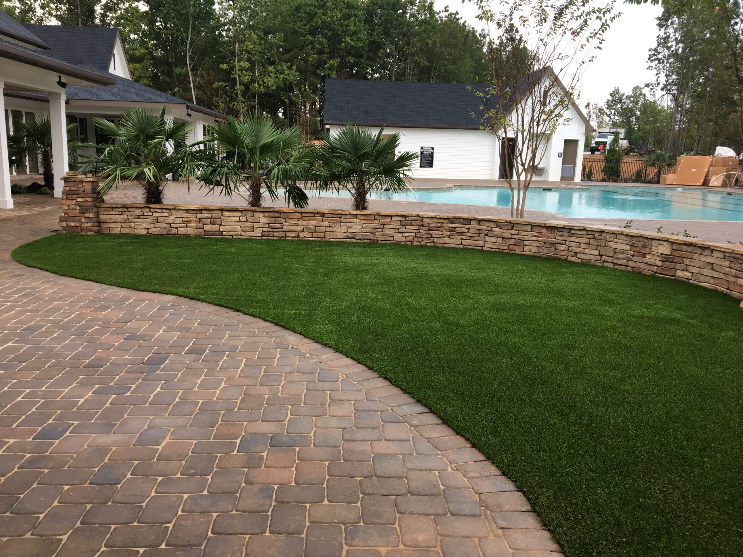 A well-manicured backyard with a curved brick patio, lush green artificial turf, a stone retaining wall, palm trees, and a clear blue swimming pool.