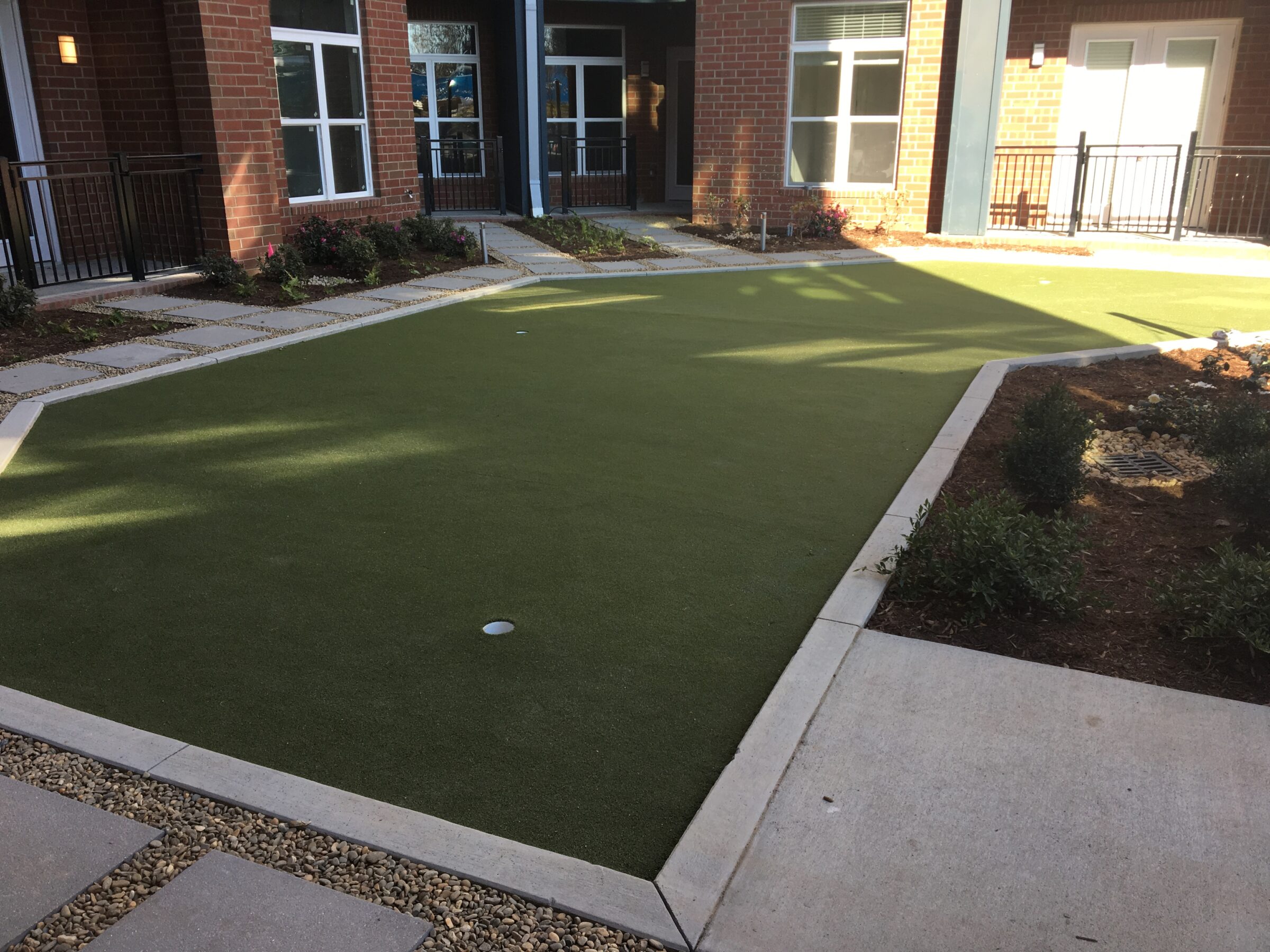 A green artificial turf putting green is surrounded by walkways, flower beds, buildings with brick walls, and metal railings under a clear sky.