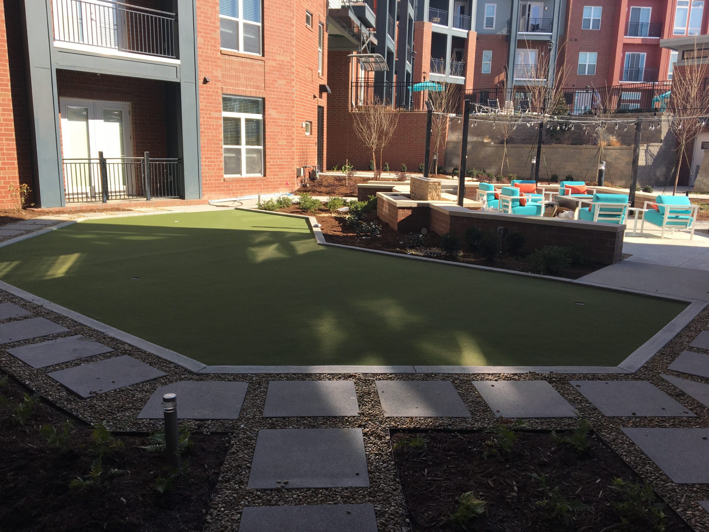 This image depicts a modern apartment courtyard with artificial turf, concrete pavers, outdoor furniture, young trees, and a string light setup over a seating area.